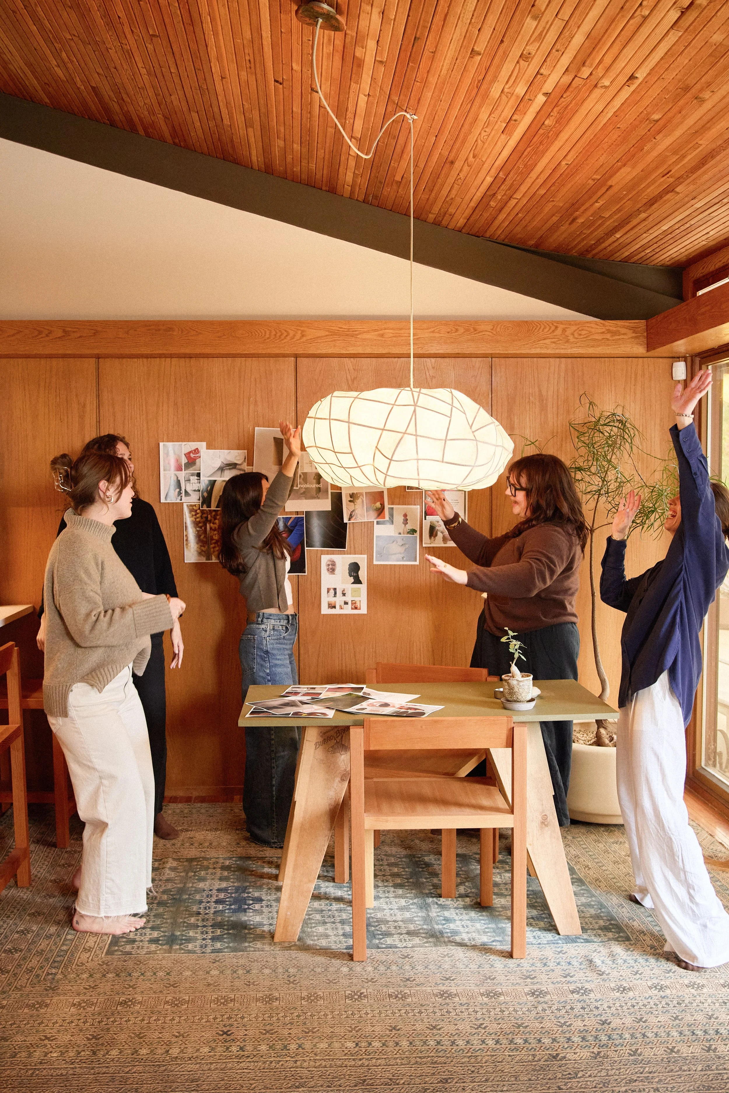 A group of five women in a room with wooden walls and ceiling, standing around a table.
Some women are raising their hands, and they seem to be engaging in a discussion or activity together.
There are photographs and images on the wall behind them and some papers on the table.