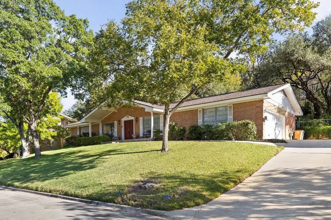 A suburban single-story brick house with a front porch, surrounded by lush green trees and a well-maintained lawn, with a driveway on the right side.