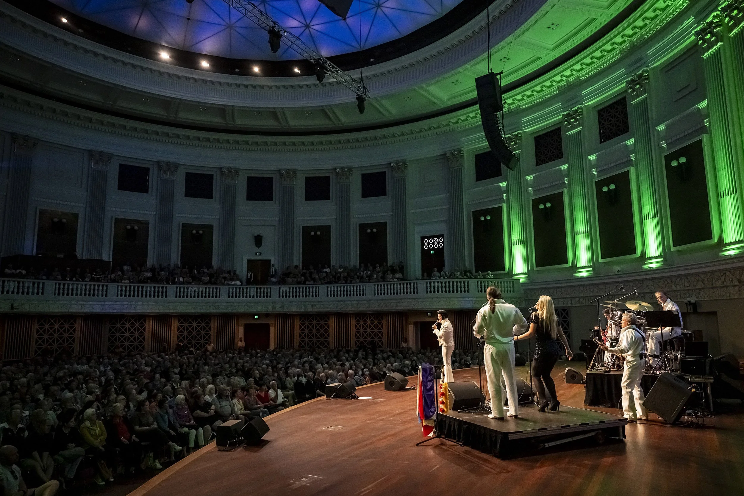 A large indoor concert venue with arched ceiling and green-lit columns, featuring a band performing on stage in front of a full audience. The band members are playing instruments and singing.
