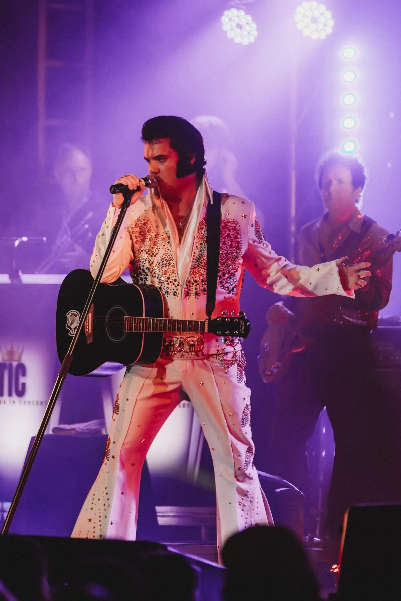 Elvis Presley impersonator singing into a microphone, playing guitar, dressed in a white jumpsuit with red and gold embellishments, performing on stage with backup musicians and purple stage lighting.