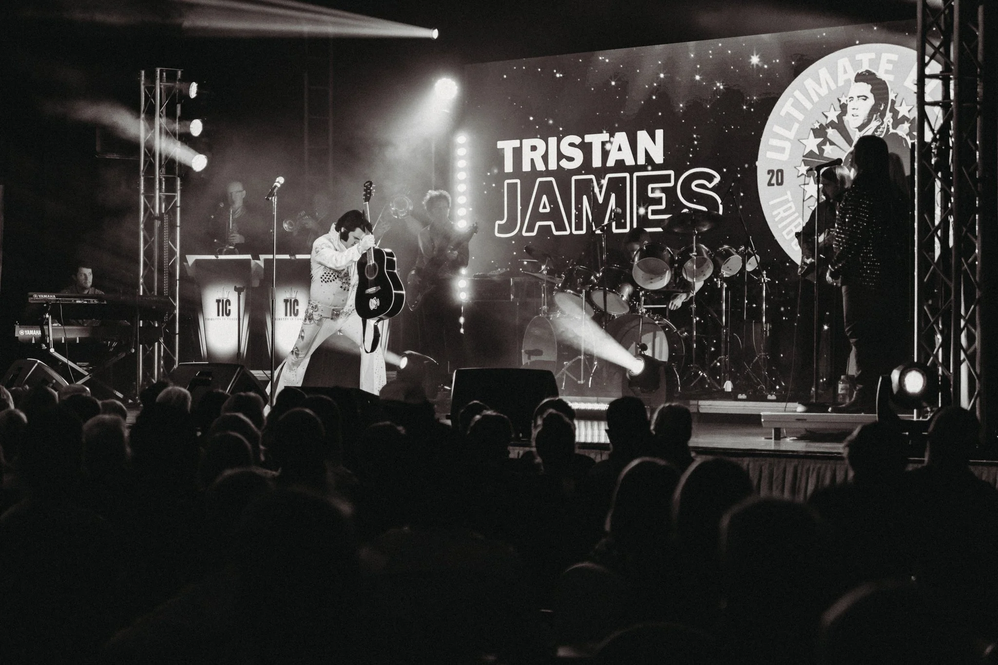 A black and white photo of a live music performance on stage with a band and a singer holding a guitar. The stage background displays the name 'Tristan James.' There is an audience watching in the foreground.