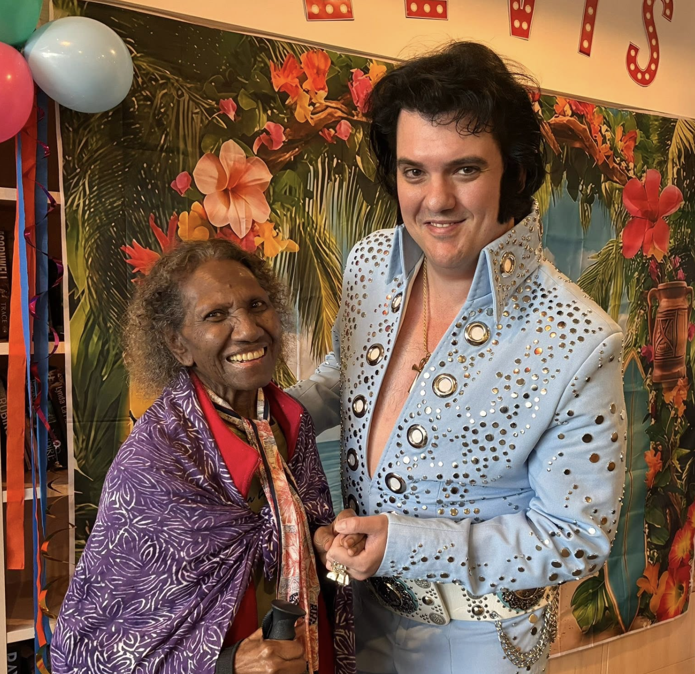 A performer dressed as Elvis Presley entertaining elderly residents at a nursing home or assisted living facility, with residents clapping and smiling, some seated in wheelchairs or with walkers, in a brightly decorated common room.