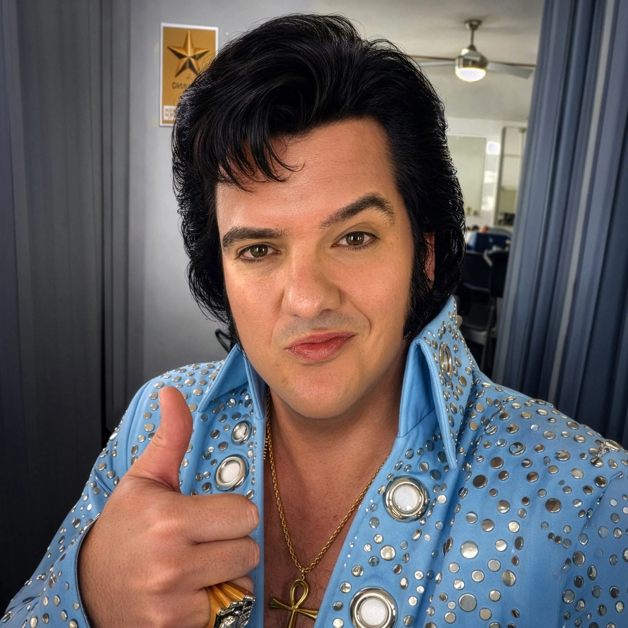 A man with dark hair and a serious expression wearing an off-white jacket with multicolored beads and fringes, standing with arms outstretched against a dark background.