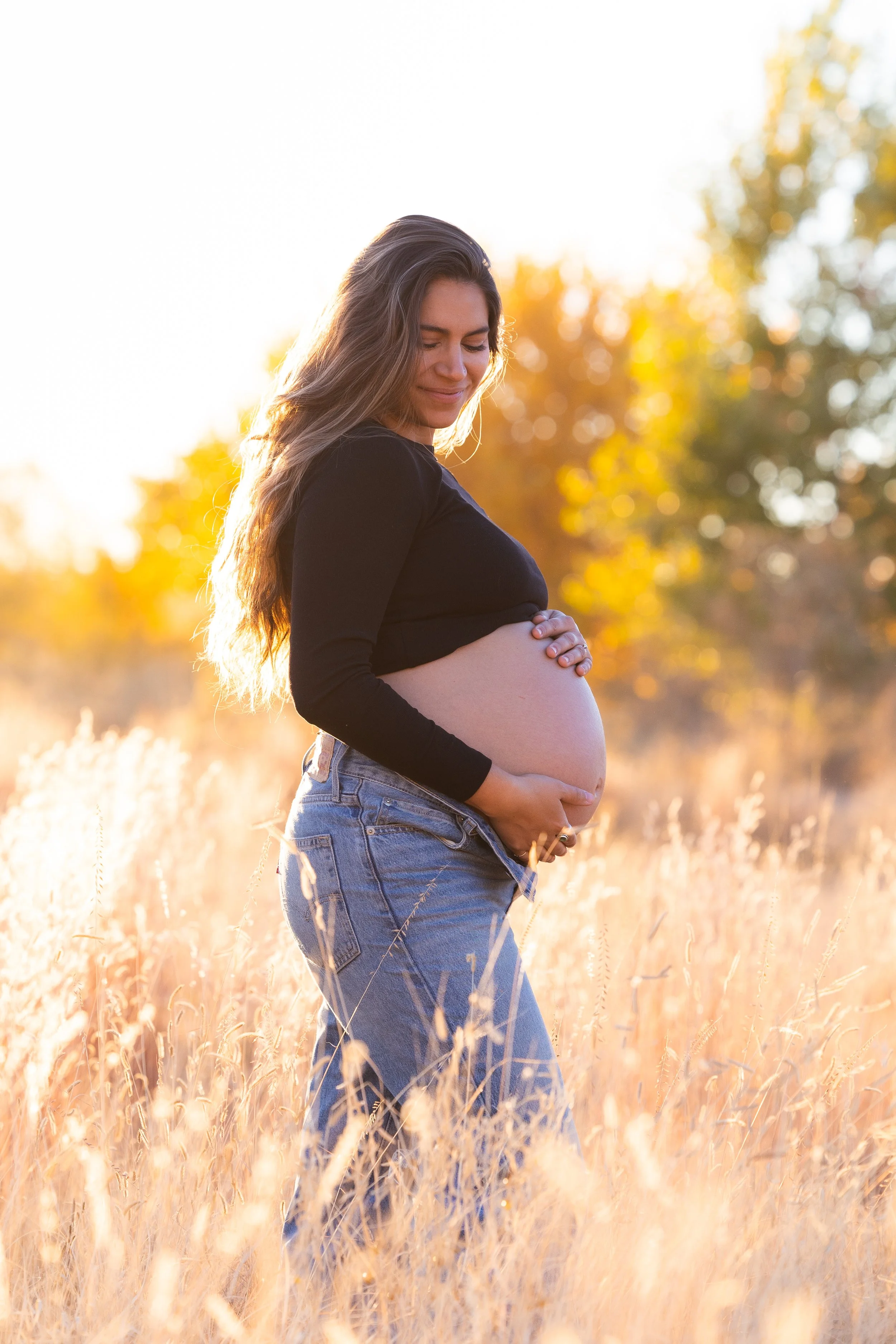 A pregnant woman standing in a field of tall, golden grass during sunset, holding her belly and looking down with a peaceful expression.