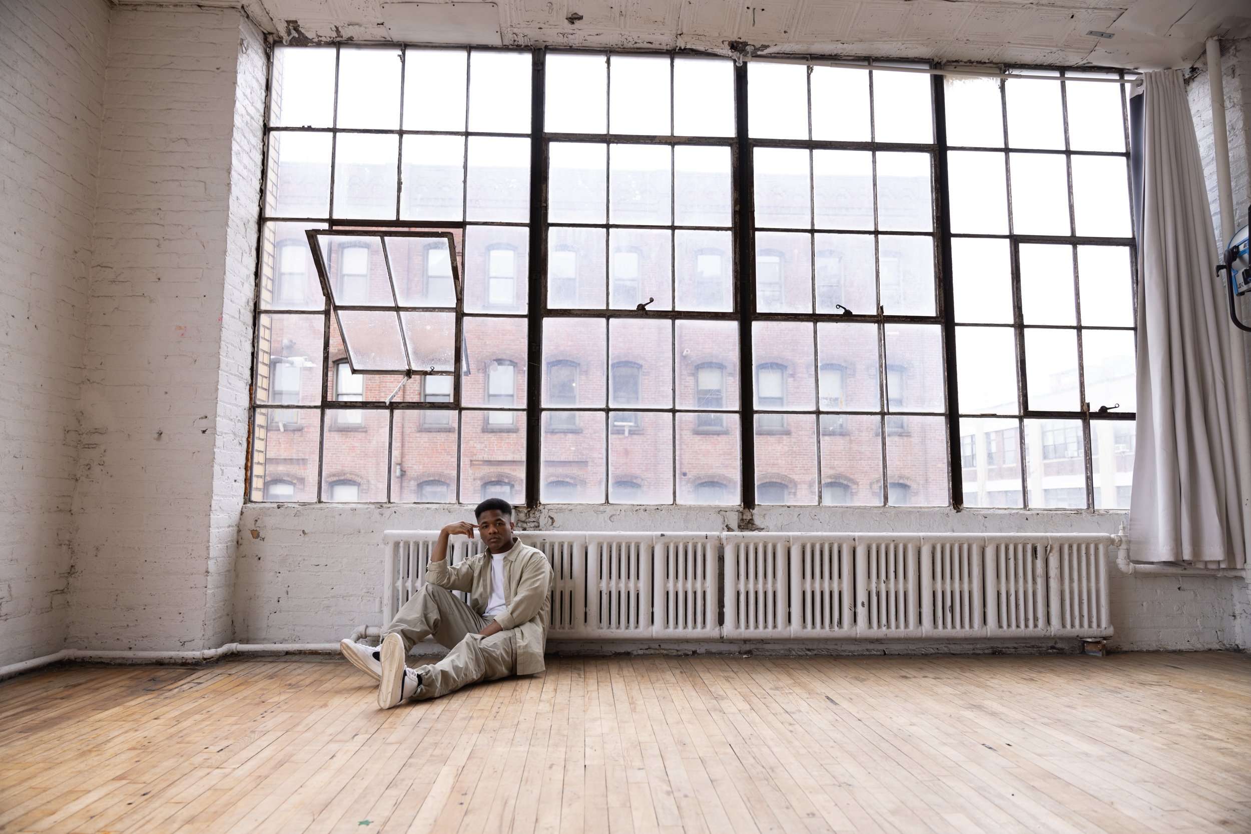 A young man in a casual beige outfit sitting on the wooden floor against a radiator, inside a spacious industrial-style room with large multi-pane windows showing a brick building outside.