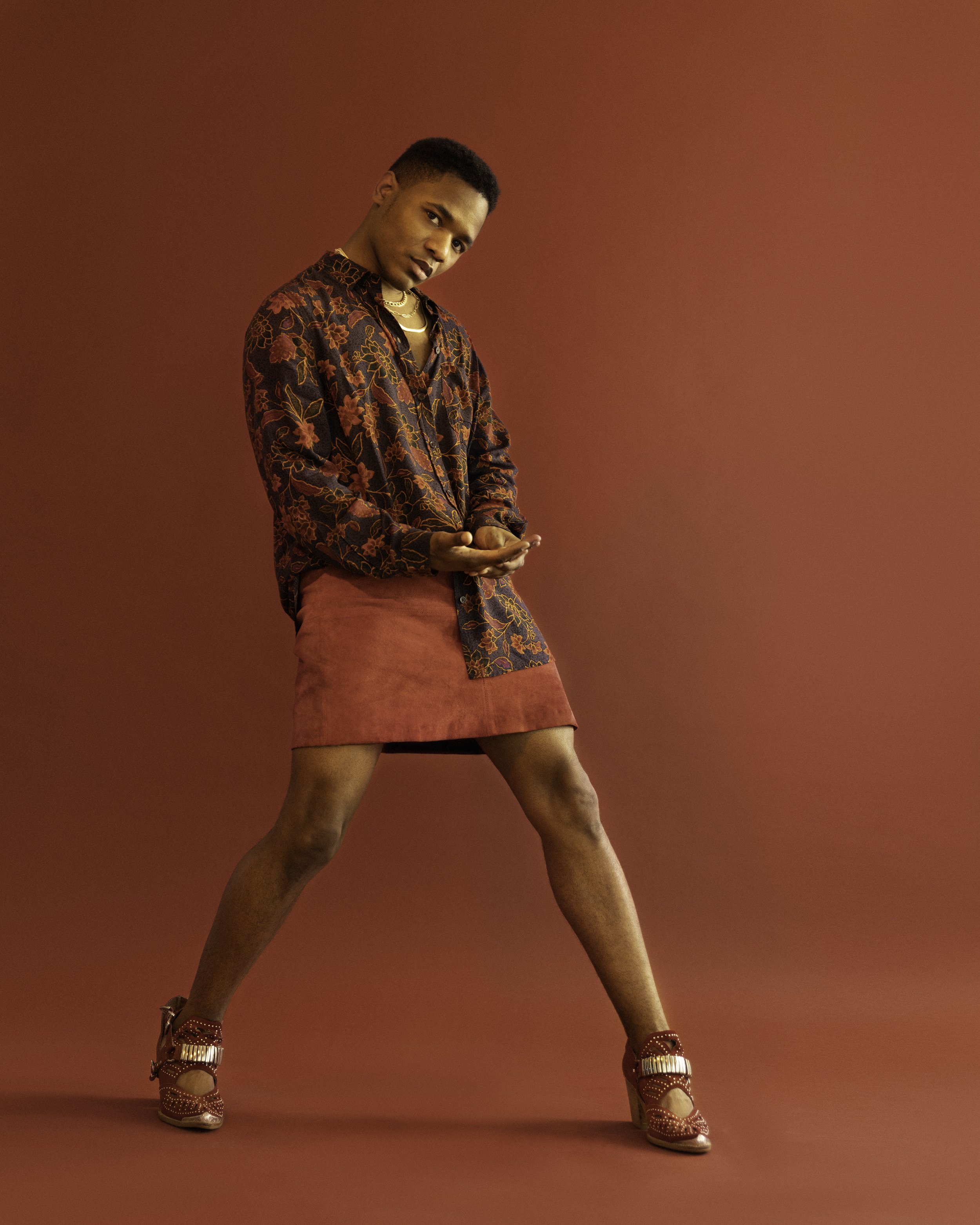 A person with dark skin and short hair posing against a plain background, wearing a patterned dark shirt, a rust-colored skirt, patterned high heels, and accessorized with layered necklaces.