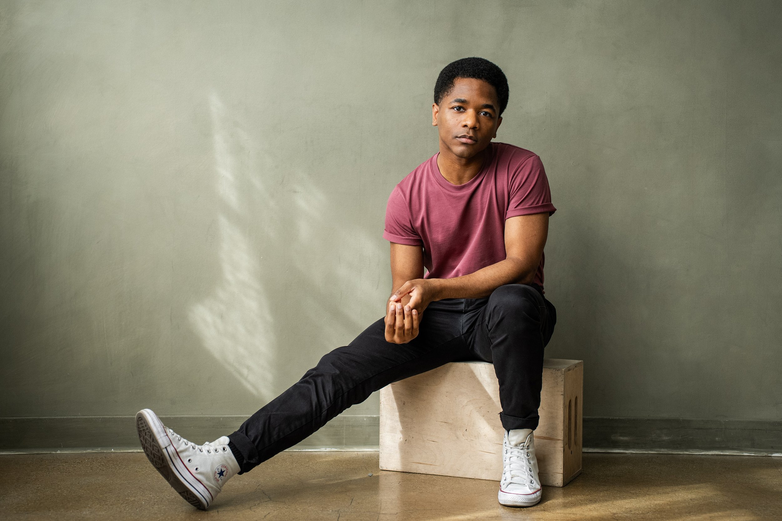 Young man sitting on a wooden box against a pale green wall, wearing a maroon t-shirt, black pants, and white sneakers, with a neutral expression.