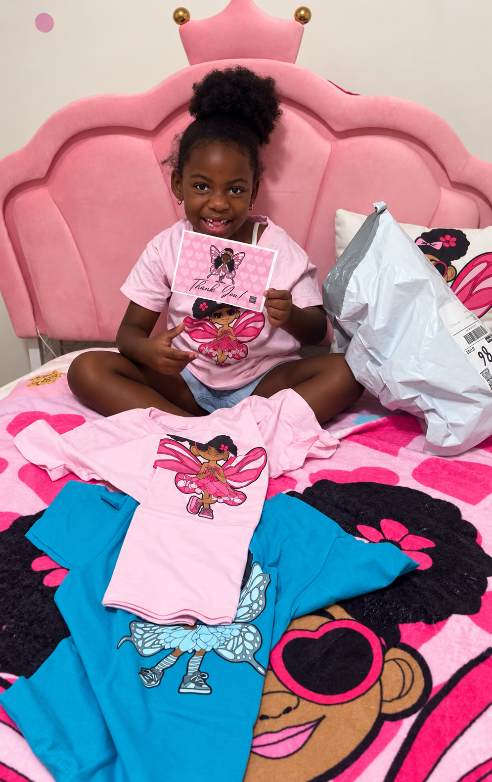 Little Black gir lin her bedroom sitting on her bed that has a Glitzy Gracelynn blanket showing her new JoJo Jollypuff and Glitzy Gracelynn shirts.
