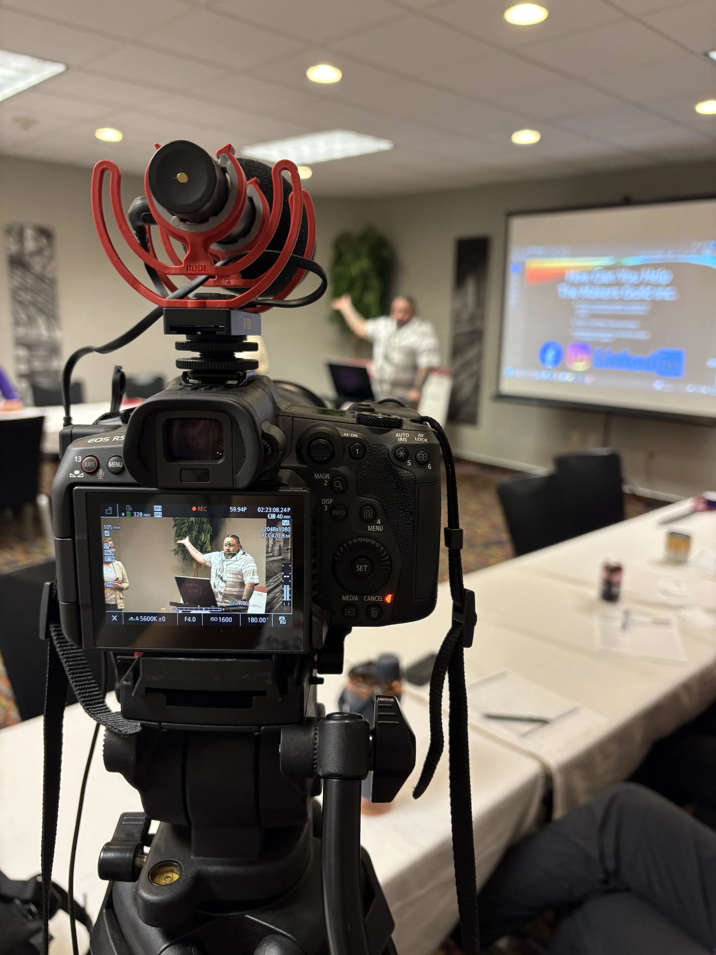 Close-up of a professional camera mounted on a tripod, capturing video of a person giving a presentation in a conference room with a projection screen and several ceiling lights.