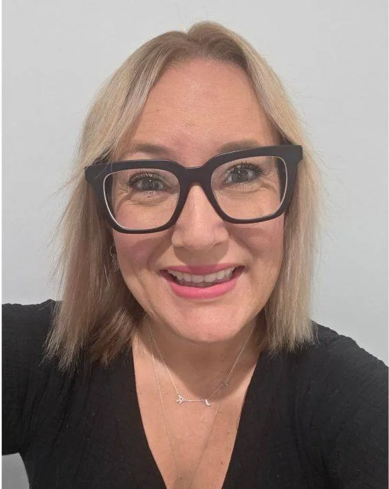 Woman with blonde hair wearing black glasses and a black top, smiling at the camera.