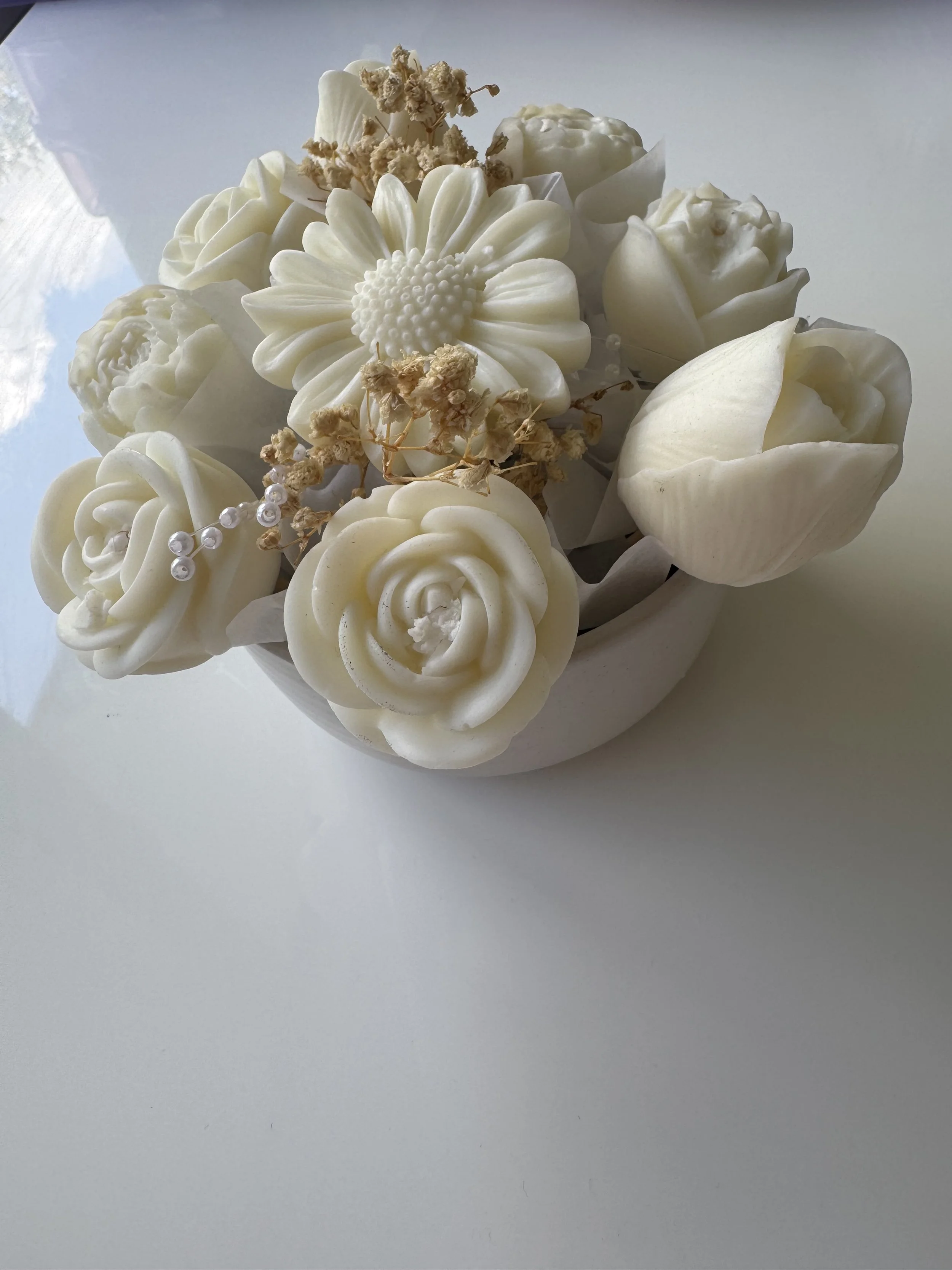 A floral arrangement of white roses, a white daisy, and small dried flowers with pearl-like beads, all in a white container.