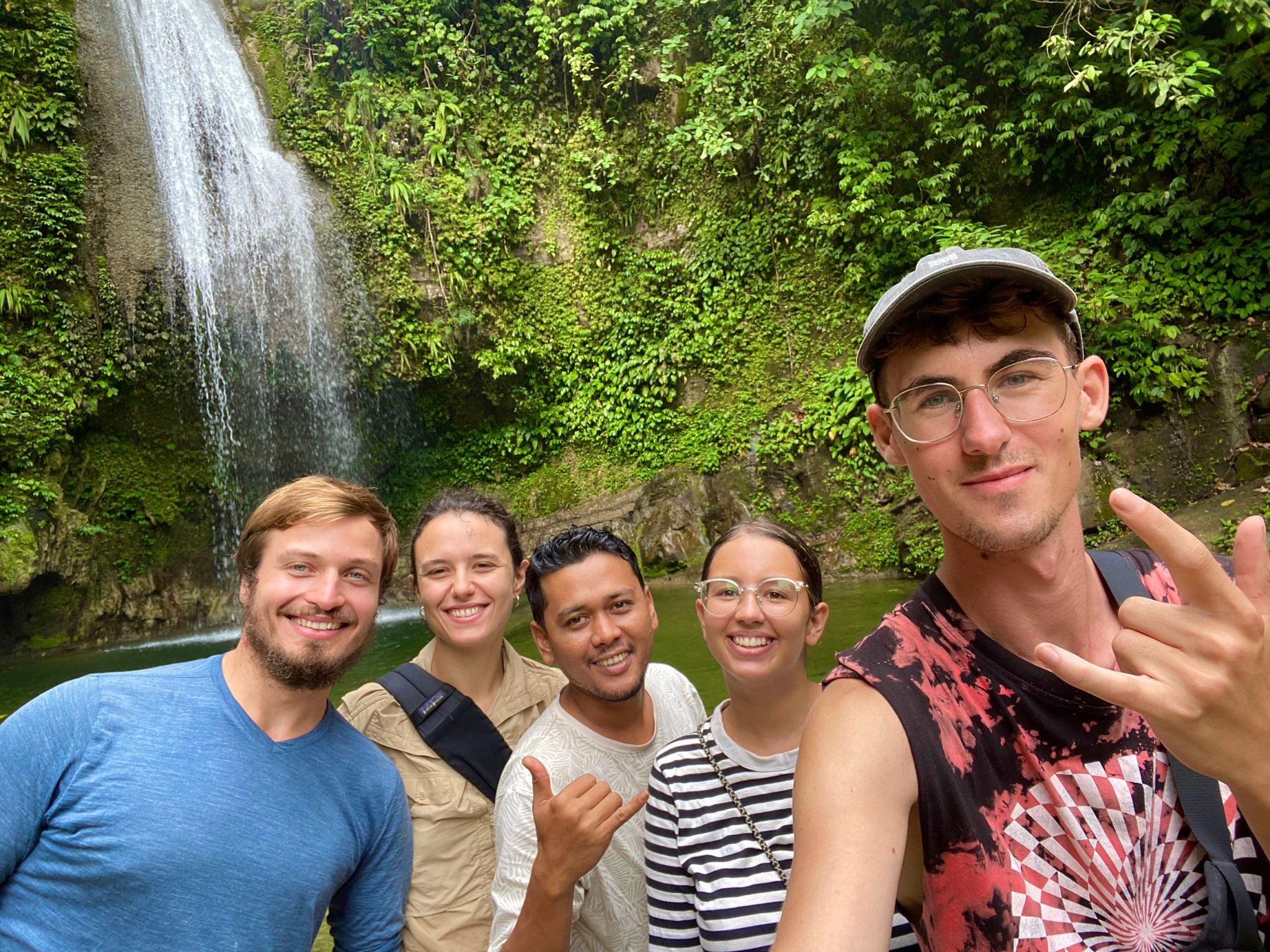Group of five friends smiling and taking a selfie in front of a waterfall surrounded by lush greenery.