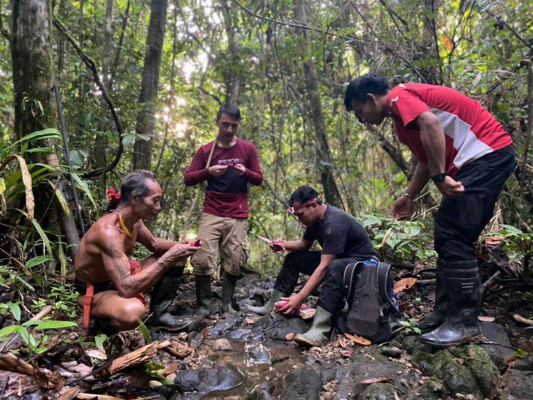Four men in a dense rainforest, two are standing and two are crouching near a small stream, with one man dressed traditionally without a shirt, and all are wearing rubber boots. They appear to be documenting or examining something in the water with s