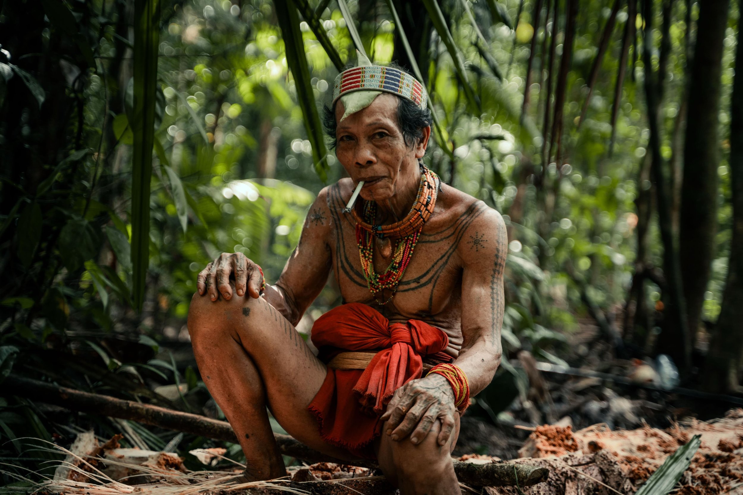An elder indigenous man sitting on the forest floor, smoking a cigarette, wearing traditional beads, tattoos, and a head covering, surrounded by dense jungle foliage.