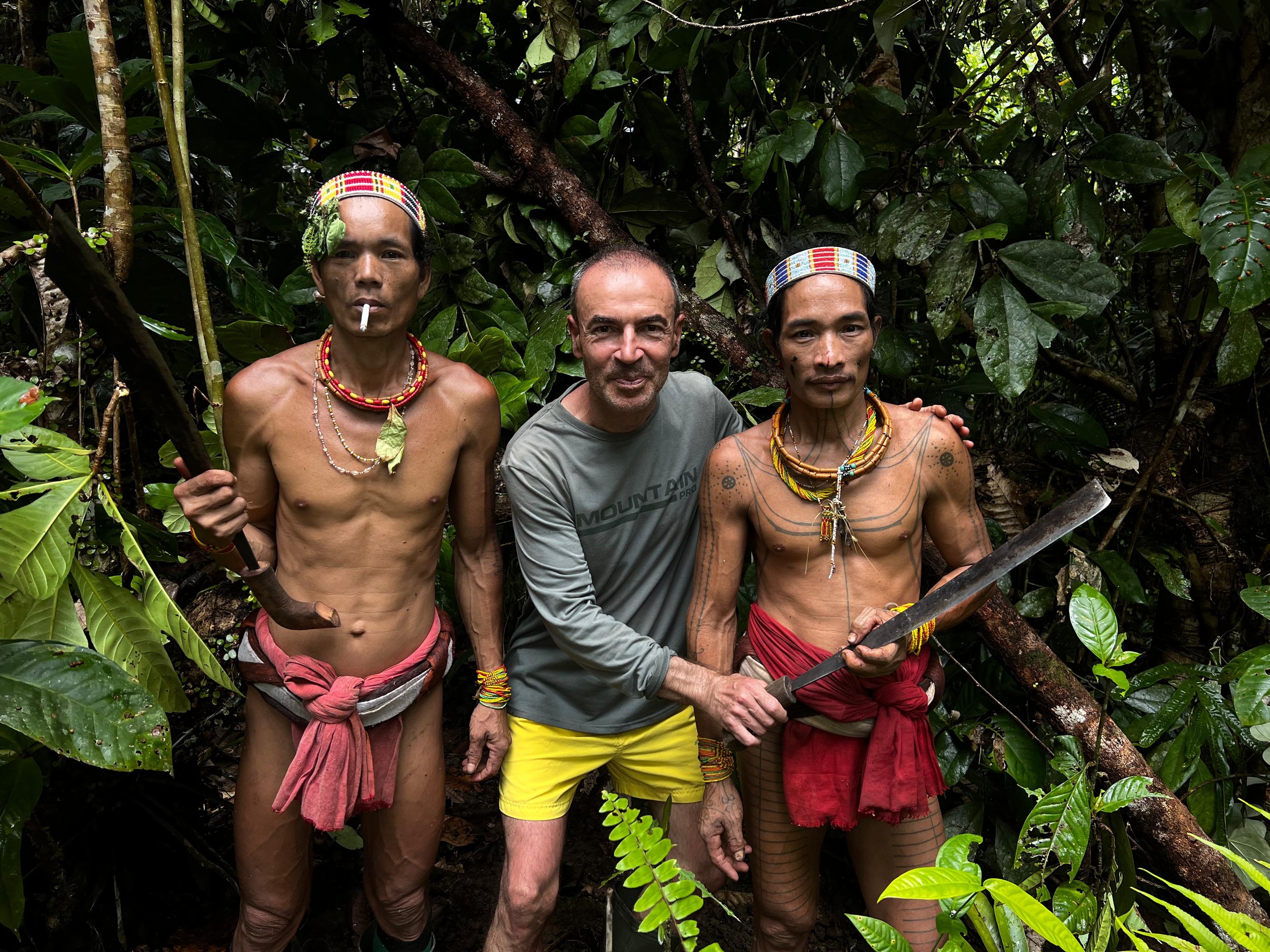 Three men standing in a dense jungle, two of whom are indigenous with traditional clothing and accessories, and one man in casual clothing.