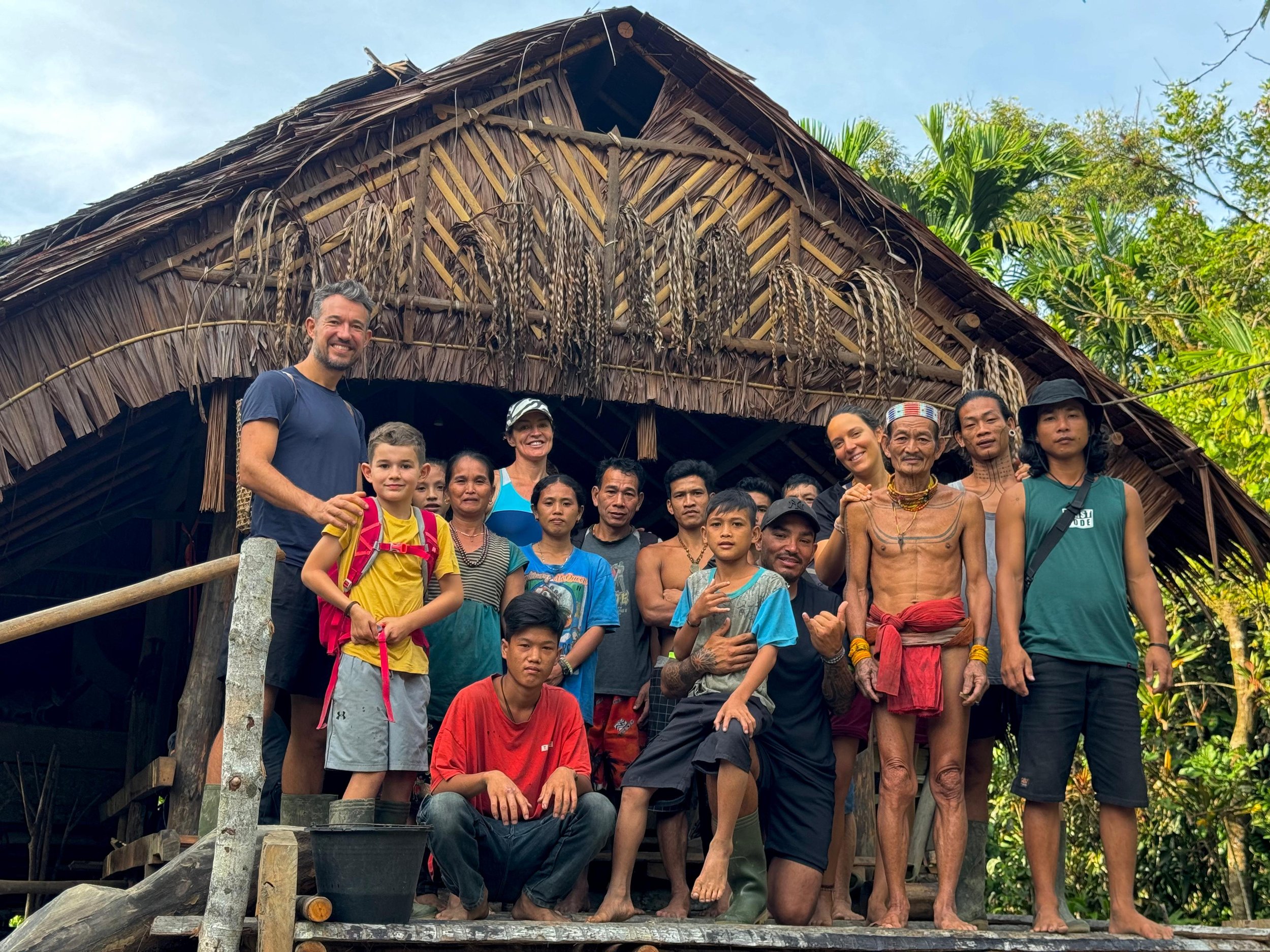 Group of tourists and the Mentawai shaman standing in front of traditional house surrounded by lush greenery.