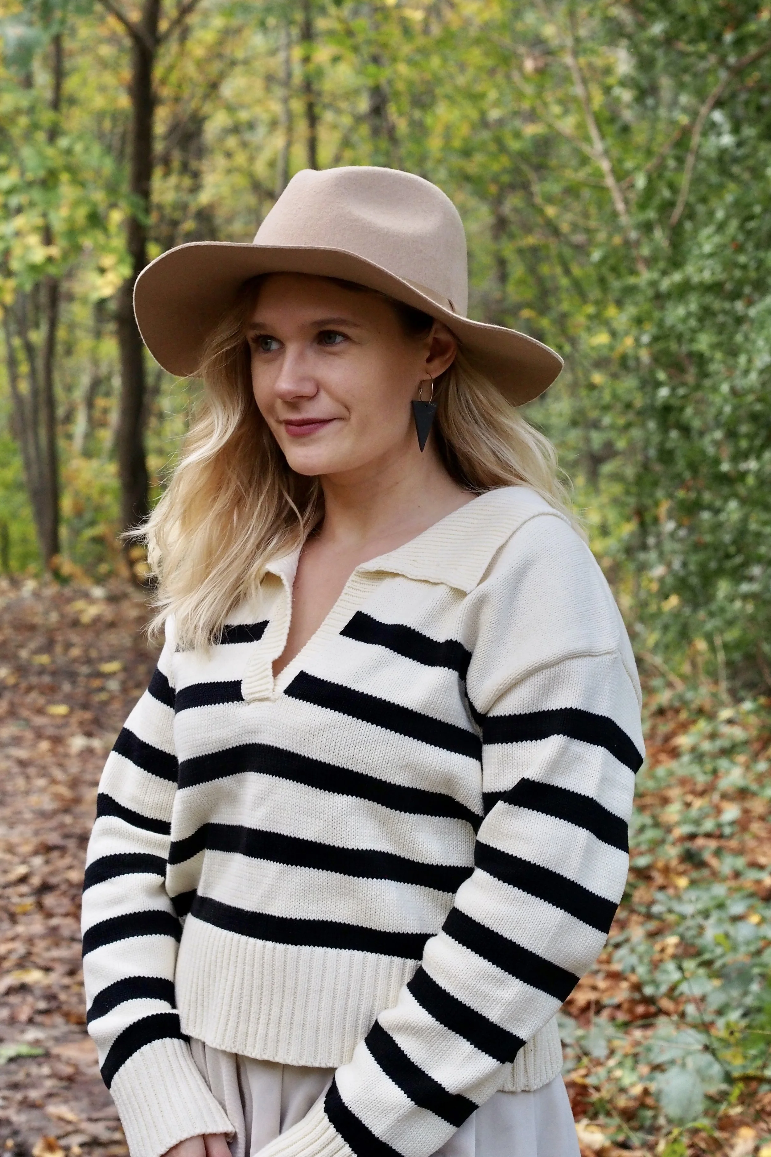 A woman with wavy blonde hair, wearing a beige wide-brimmed hat, black and beige striped sweater, and beige pants, standing outdoors in a wooded area with green and yellow leaves.