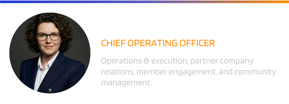 A woman with curly hair, glasses, and a dark blazer poses for a professional portrait in front of a blue background.