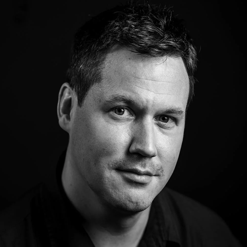 A black and white portrait of a man with short hair, wearing a dark shirt, looking towards the camera.