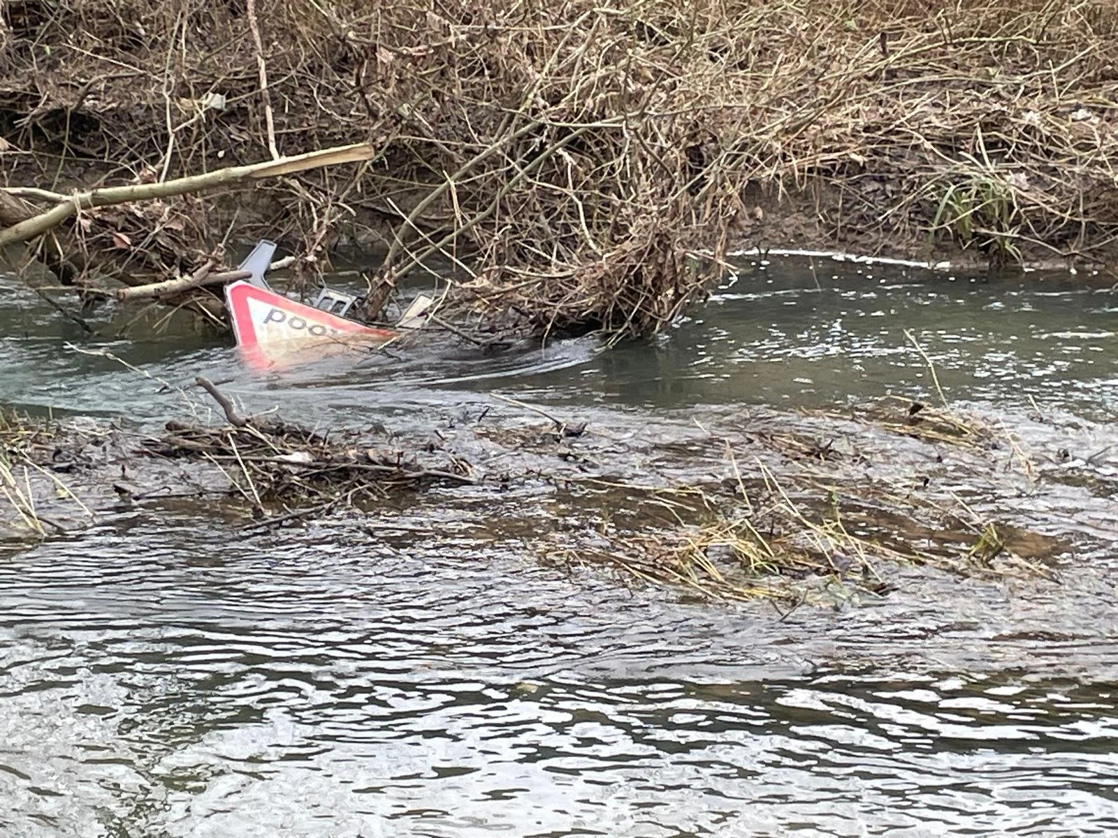 Partial view of a partially submerged damaged road sign in a creek with branches and water surrounding it.