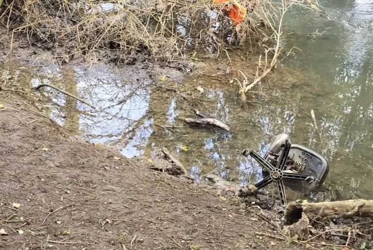 An office chair partially submerged and overturned in muddy water near a wooded shore.