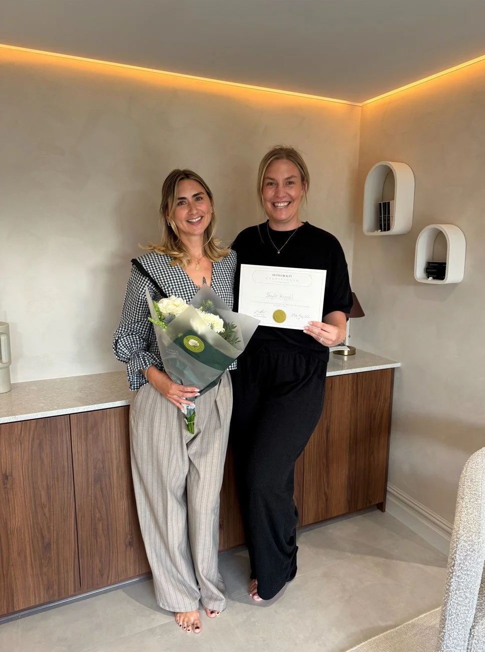 Two women standing indoors, smiling. One woman holds a bouquet of white roses and the other holds a certificate.