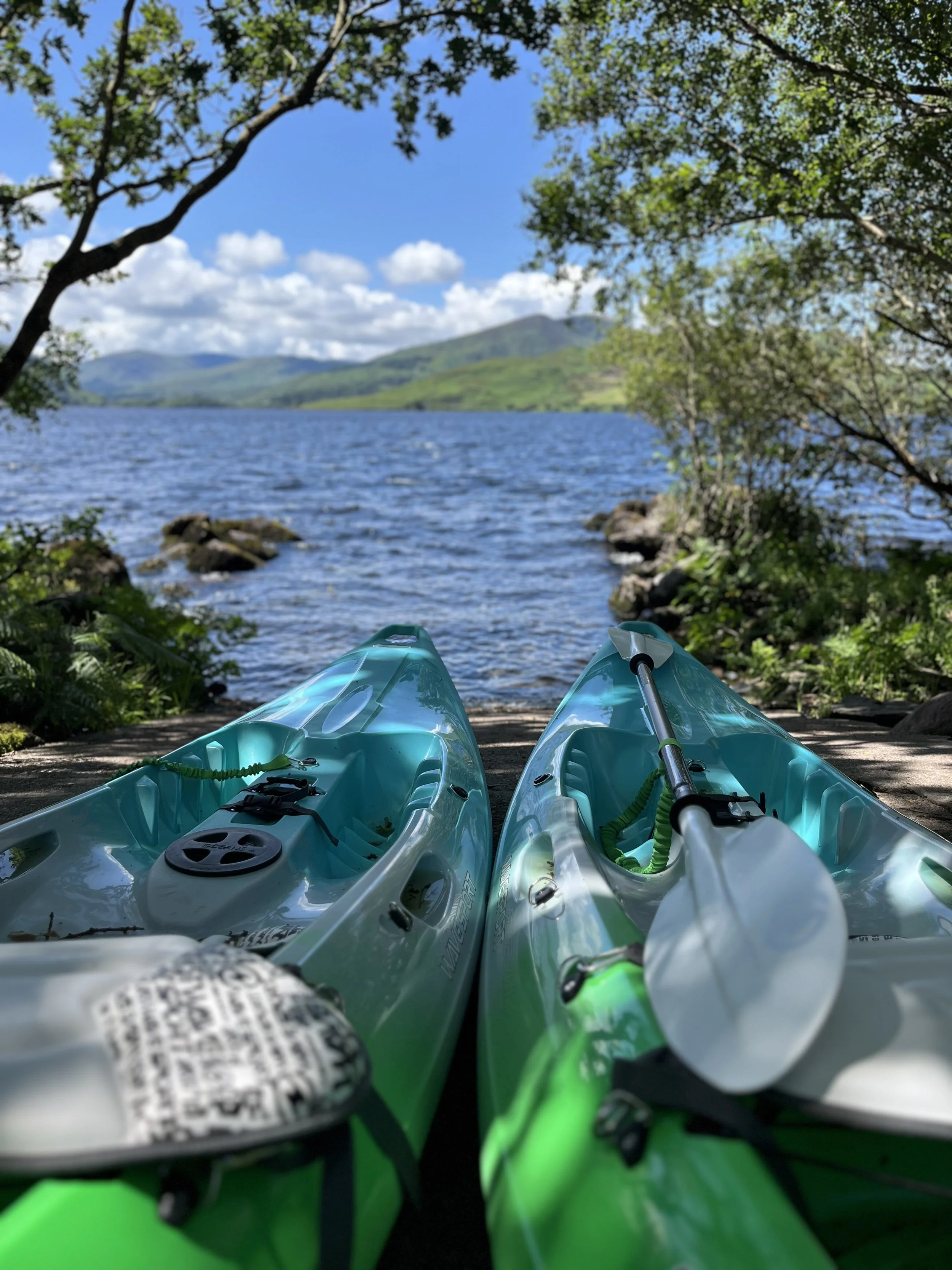 Two green kayaks on a lakeshore under trees, with a view of a lake, hills, and a partly cloudy sky in the background.
