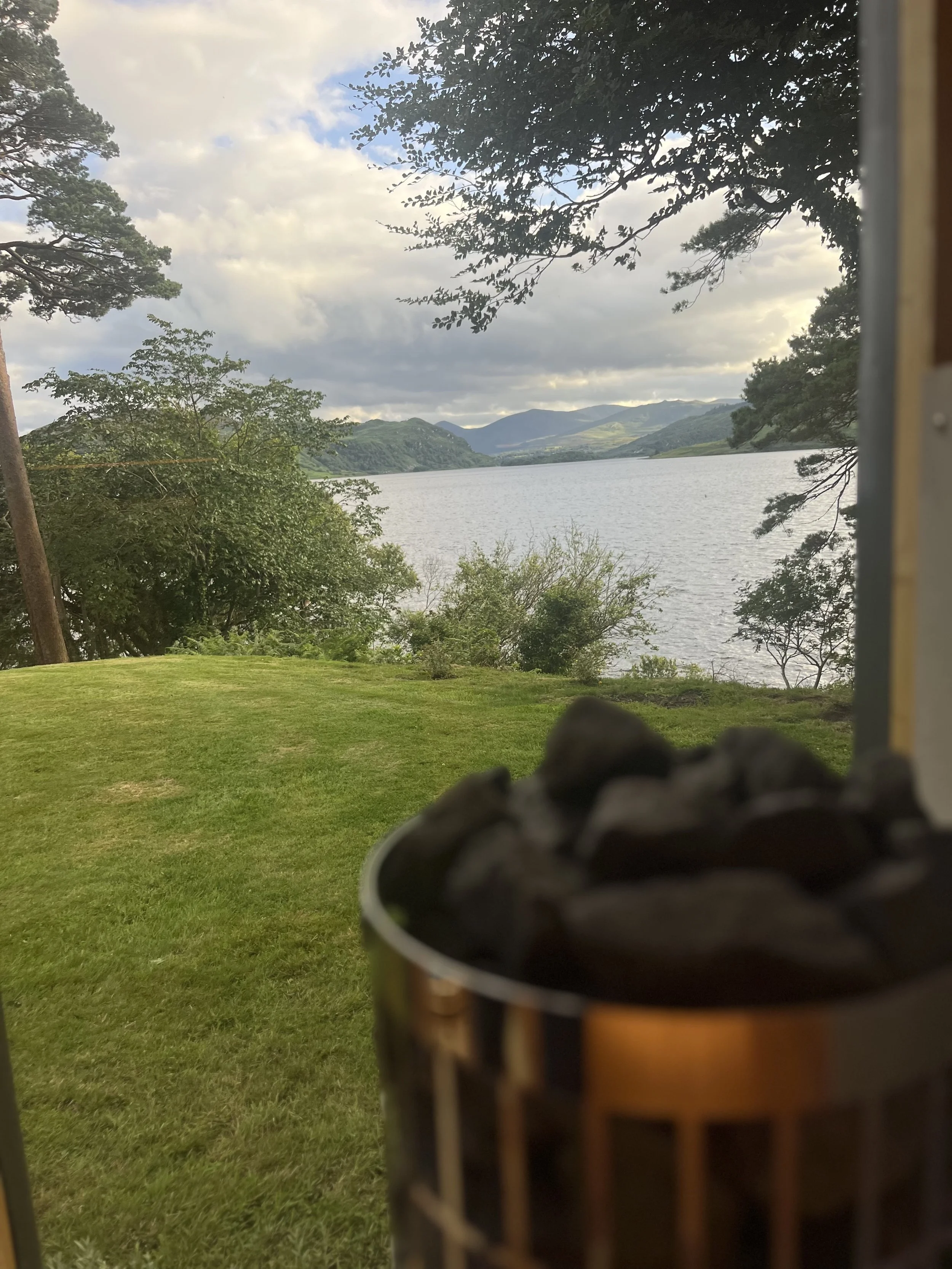 A scenic view through a window showing a lake surrounded by green hills and trees with a cloudy sky. In the foreground, a fire pit with hot coals is visible.