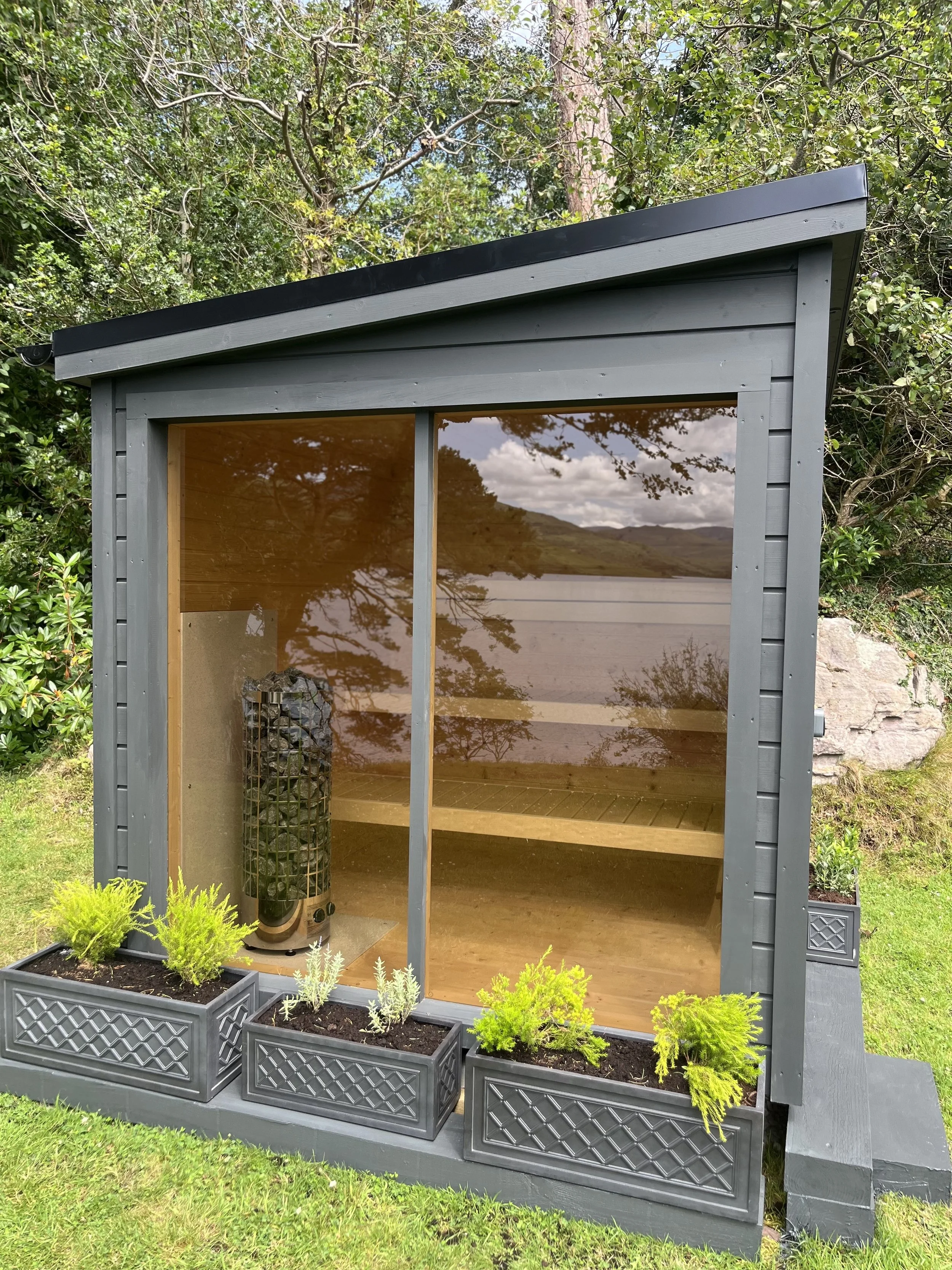Small outdoor wooden shed with gray paint and a slanted roof, surrounded by greenery and shrubs. Inside, a heater is visible through the open glass sliding door, with two wooden shelves inside. The shed is decorated with flowerpots containing various