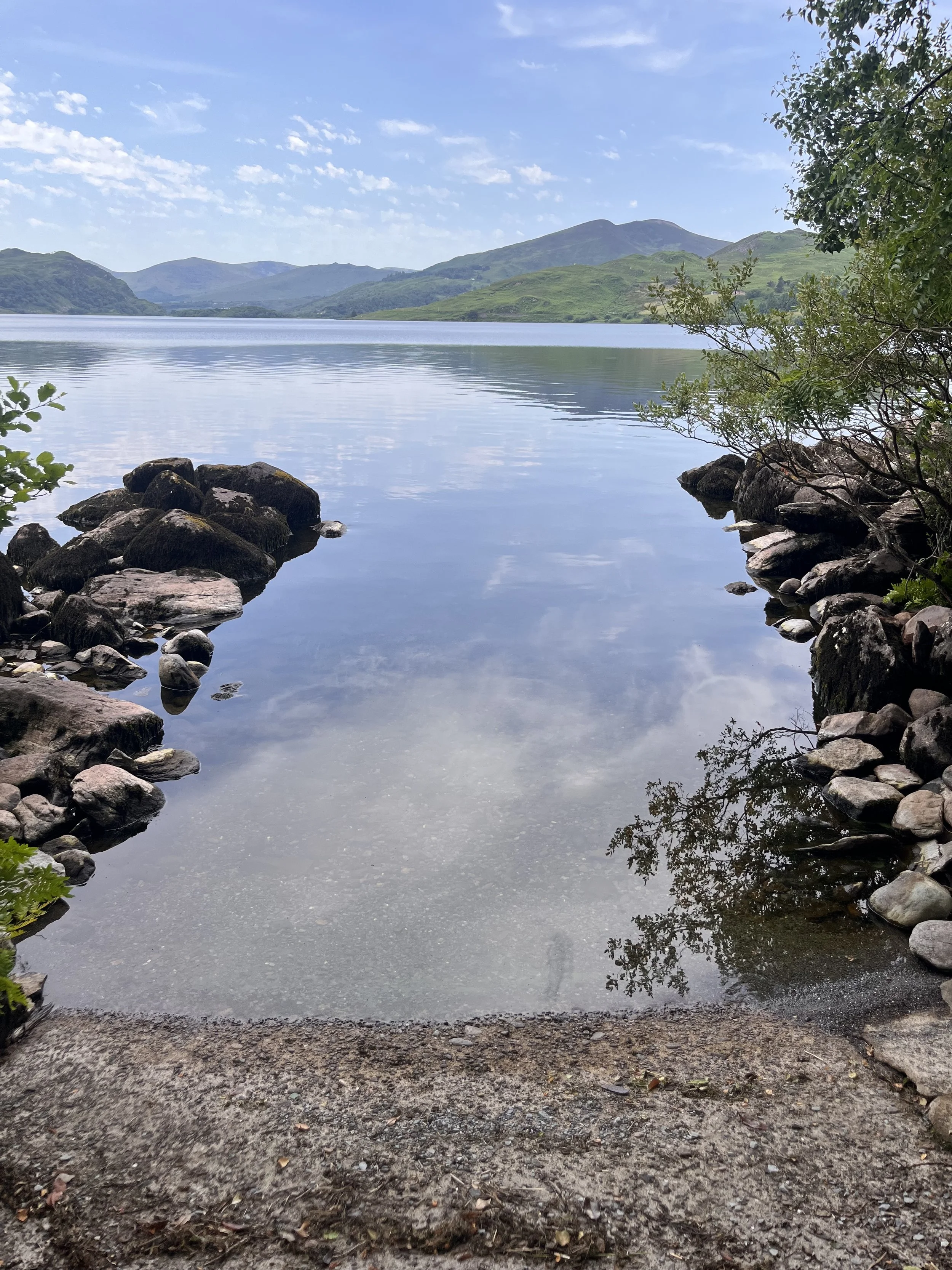 A serene lake with clear water reflecting the blue sky and scattered clouds, surrounded by green hills and mountains in the distance. Rocks and small bushes frame the shoreline.