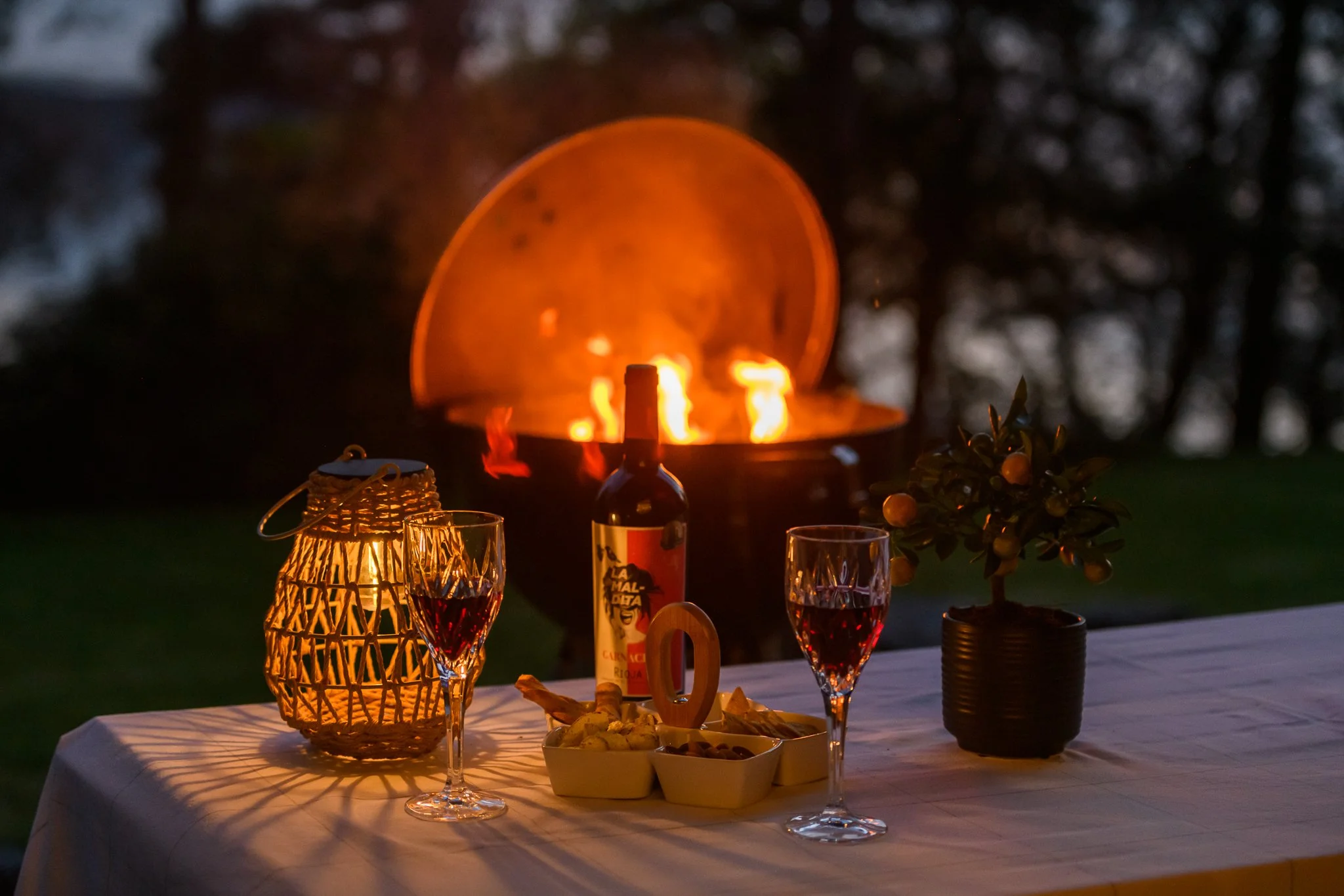 A table set for an outdoor dinner with two glasses of red wine, a bottle of wine, small bowls of snacks, a decorative lantern, and a small potted plant. In the background, a fire pit is burning with flames and sparks, creating a warm and cozy atmosph