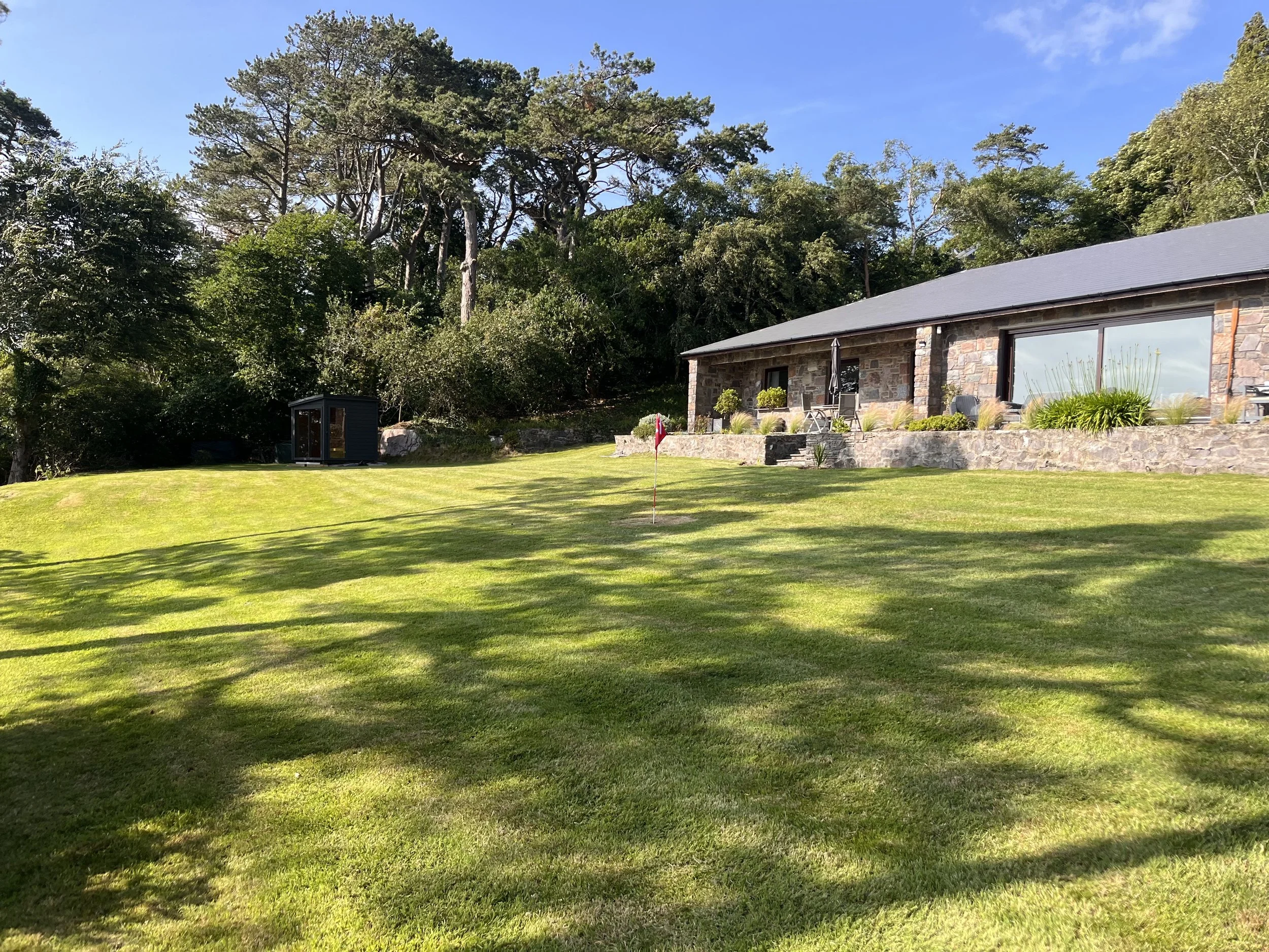 A house with a stone exterior and large windows is situated on a grassy lawn with a small golf hole and flag right in front. Trees surround the property, and the sky is clear and blue.