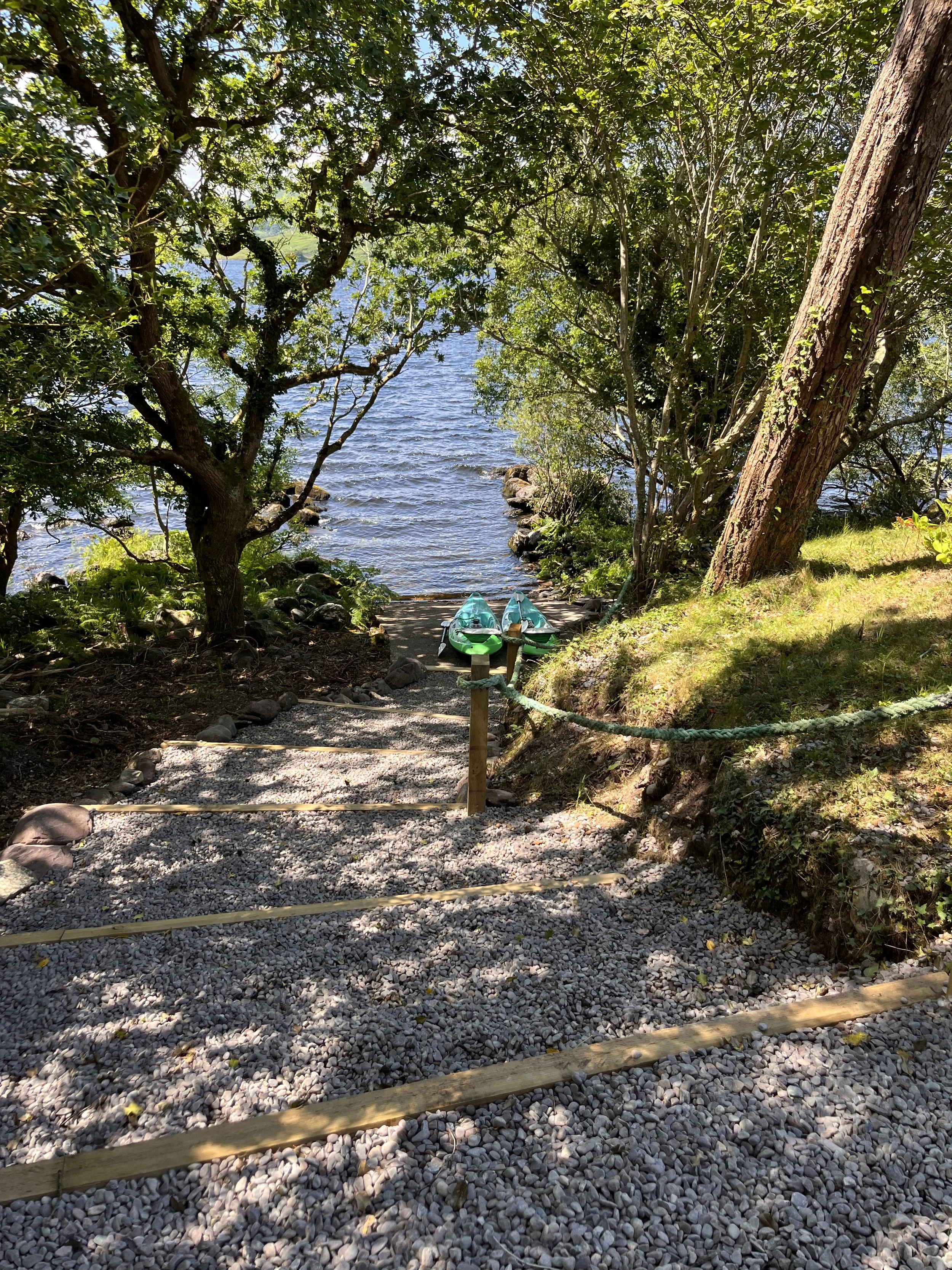 A gravel trail leading down to a lake, with two kayaks near the shoreline, surrounded by trees and greenery.