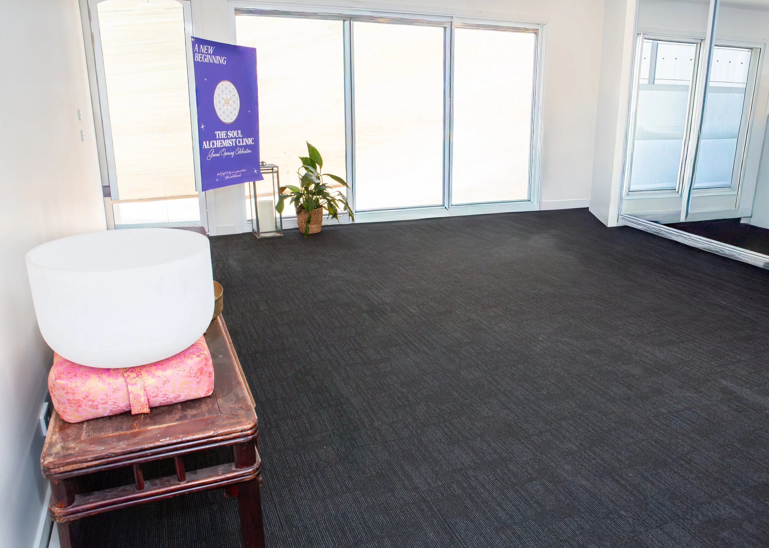 Empty room with dark carpet, large windows, a potted plant, a poster with blue background and white text, and a decorative bowl on a wooden table.