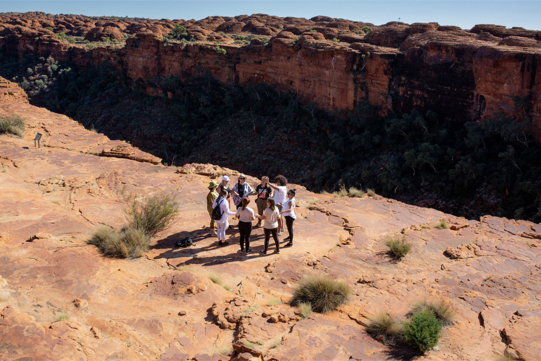 Group of people on a rocky desert landscape with cliffs in the background, some with backpacks, taking photos and talking.