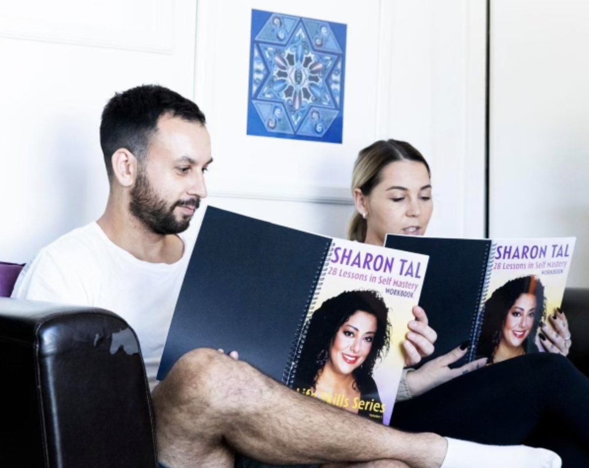 A man and a woman sitting on a couch and reading books titled 'Sharon Tal: 28 Lessons in Self Mastery Workbook.' The man has short dark hair and a beard, wears a white T-shirt, and has a skateboard beside him. The woman has light brown hair tied back and is also reading the book. There is a blue geometric artwork on the white wall behind them.