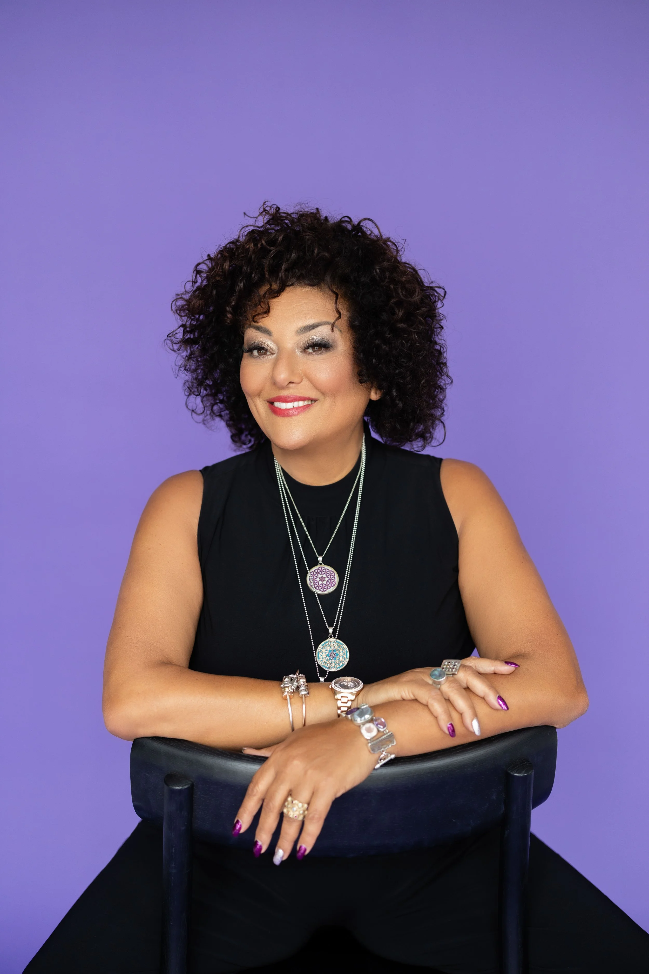 A woman with curly dark hair wearing a black sleeveless top, multiple necklaces, and various rings and bracelets, sitting on a chair with arms crossed, smiling with a purple background.