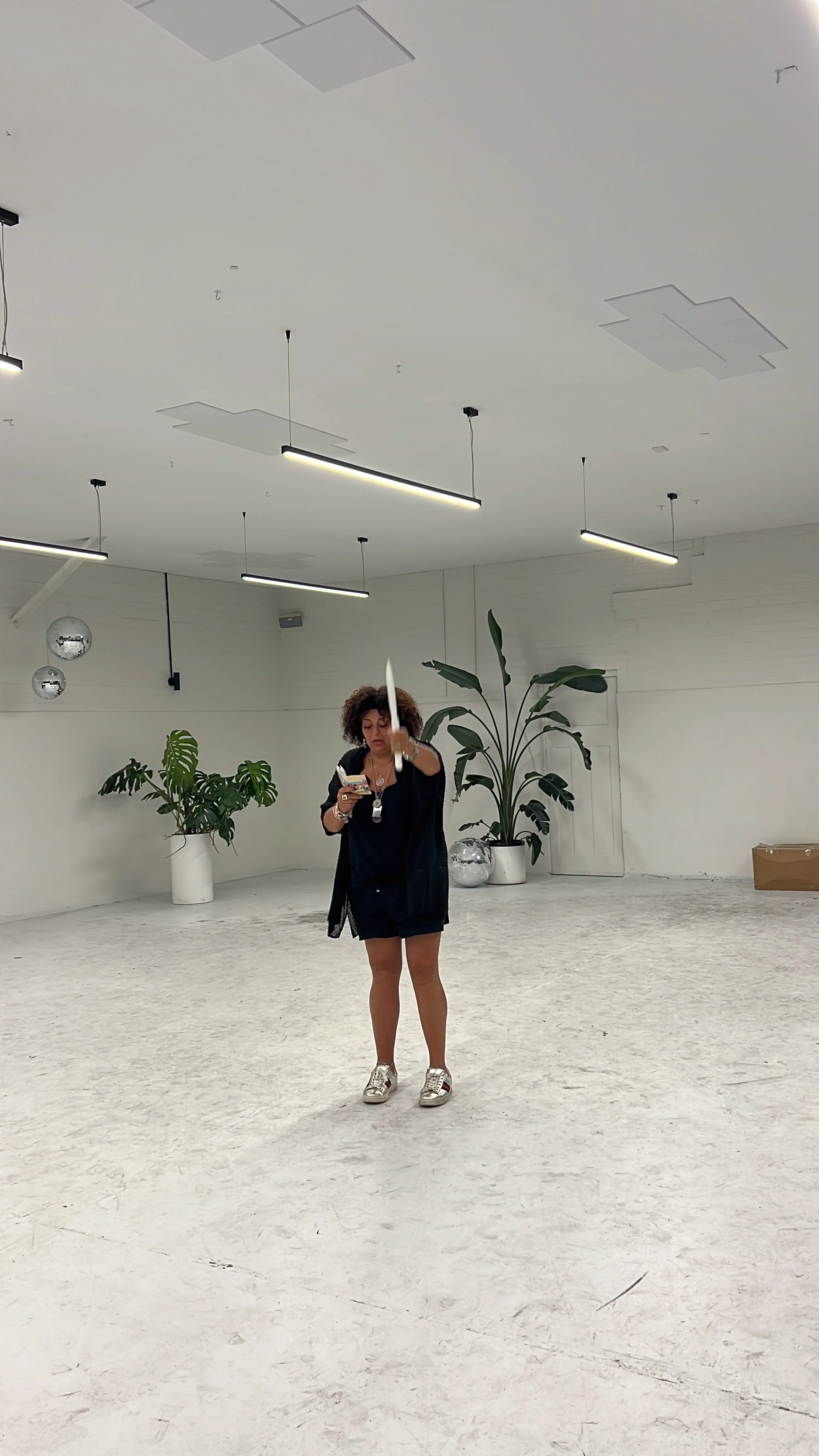 A woman dancing or moving in an empty, minimalist room with white walls and ceiling, decorated with large potted plants, metallic spheres, and suspended linear lights.