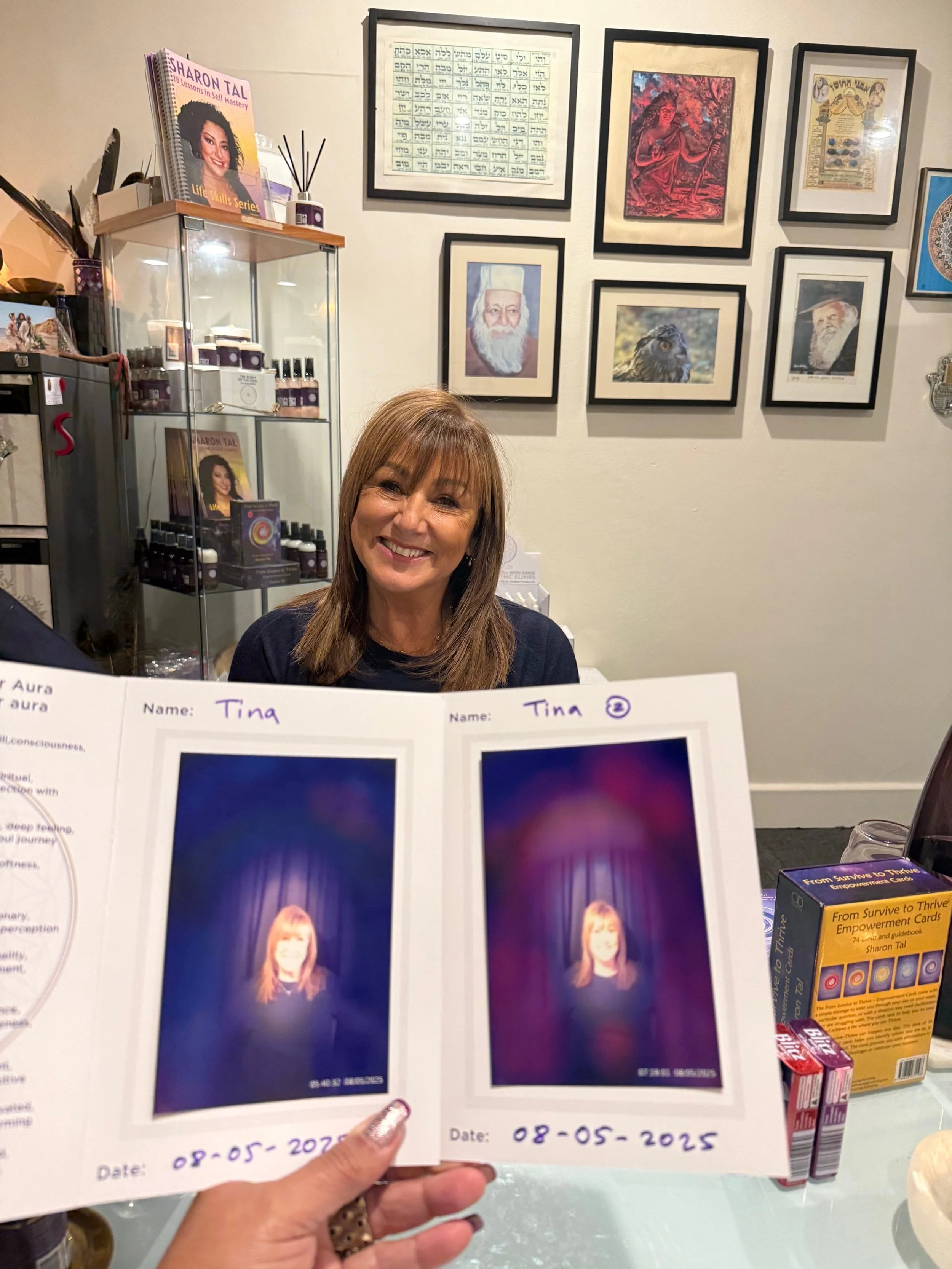 A woman is smiling while sitting at a table, with framed artwork and photographs hanging on the wall behind her. She's behind a booklet displaying two photographs of herself with the date 08-05-2025 written underneath.