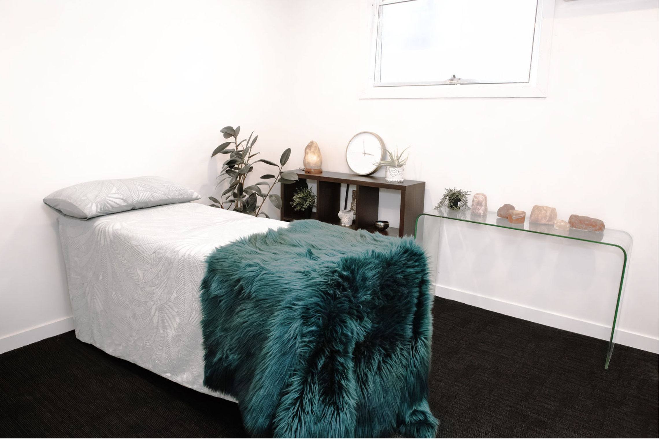 Minimalist bedroom with a bed, teal faux fur throw, wooden shelf with decor, salt lamps, plants, and a glass console table with stones.