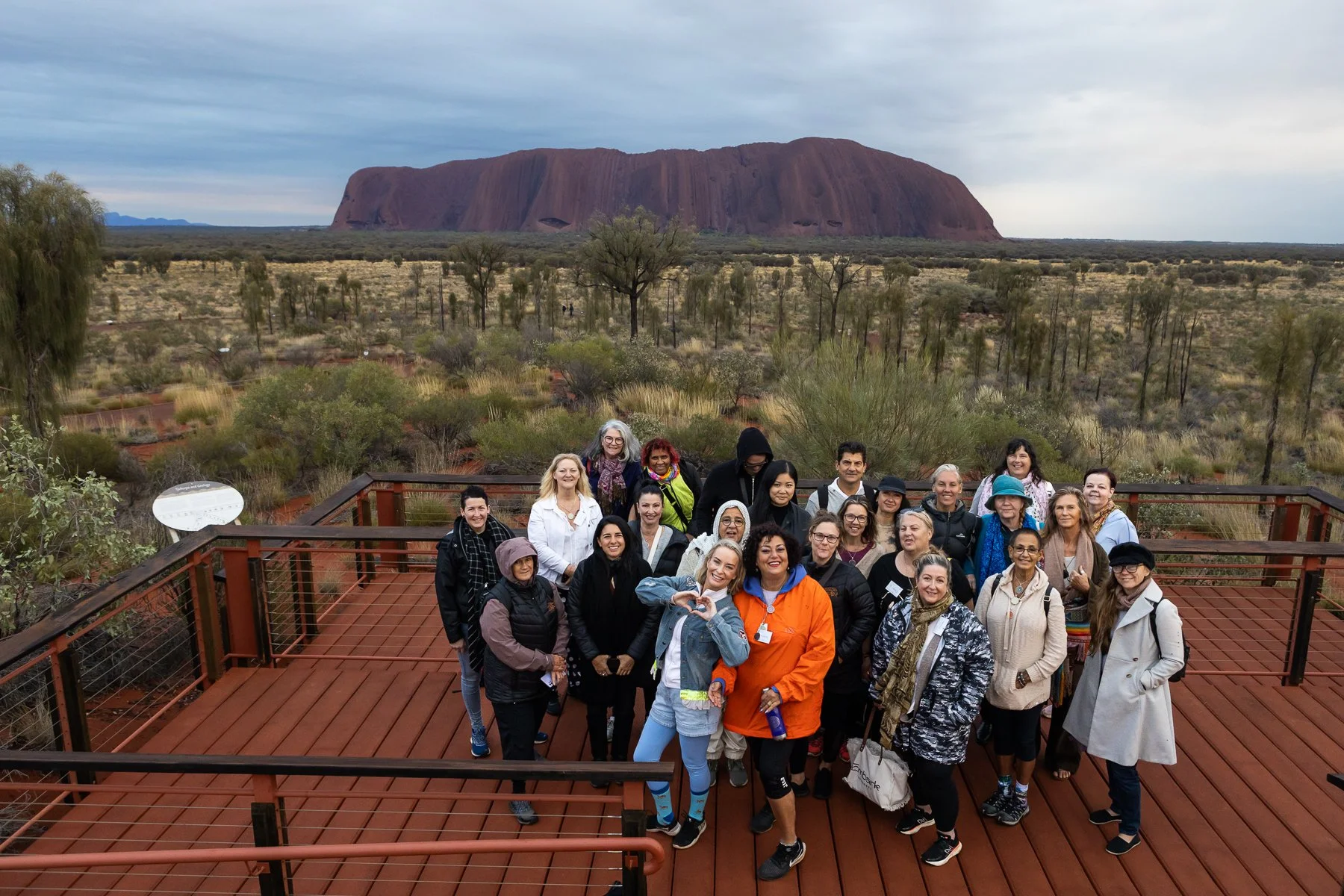 Group of people standing on a wooden observation deck with Uluru in the background, in a desert landscape with sparse vegetation and cloudy sky.
