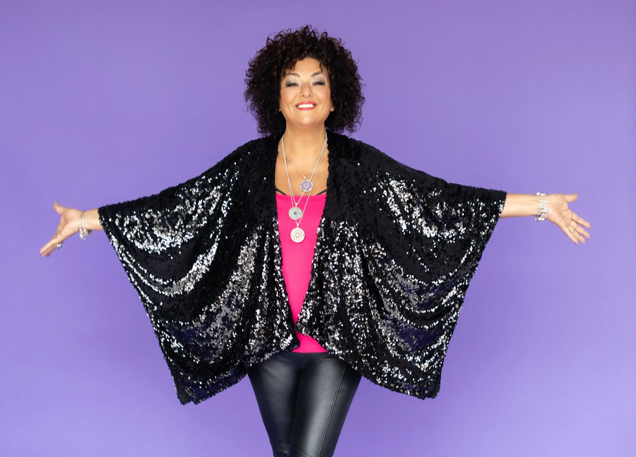 Woman with curly hair smiling and standing with arms outstretched, wearing a black sequined shawl, pink top, and black leather pants against a purple background.
