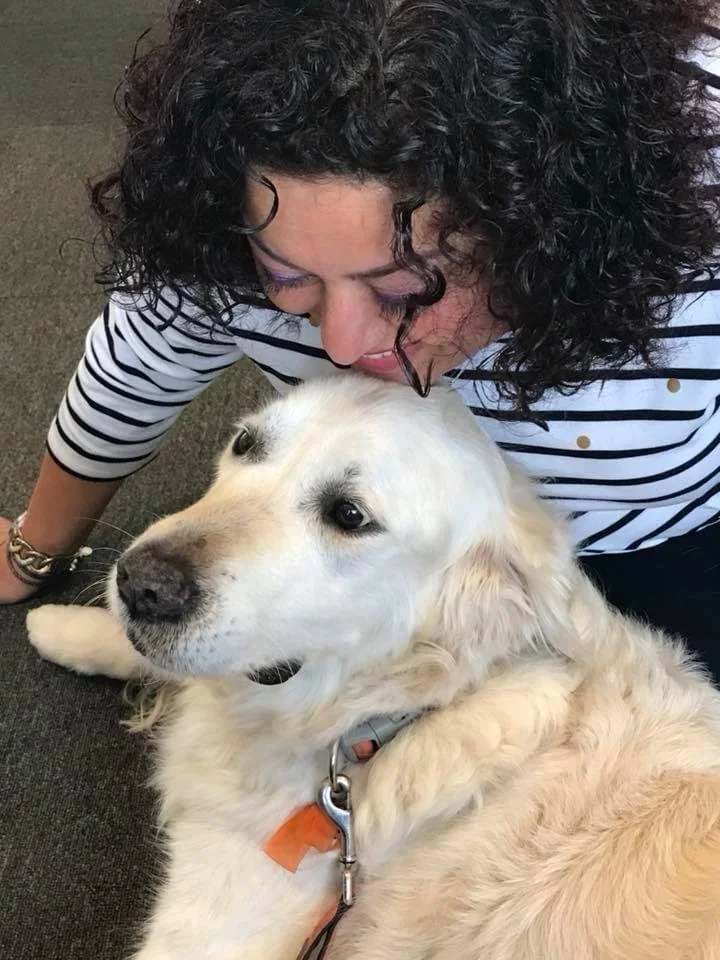 A woman with curly hair and wearing a striped shirt is lying on the floor, looking down at a large, light-colored dog lying next to her. The dog appears relaxed and is wearing a collar with an orange tag.
