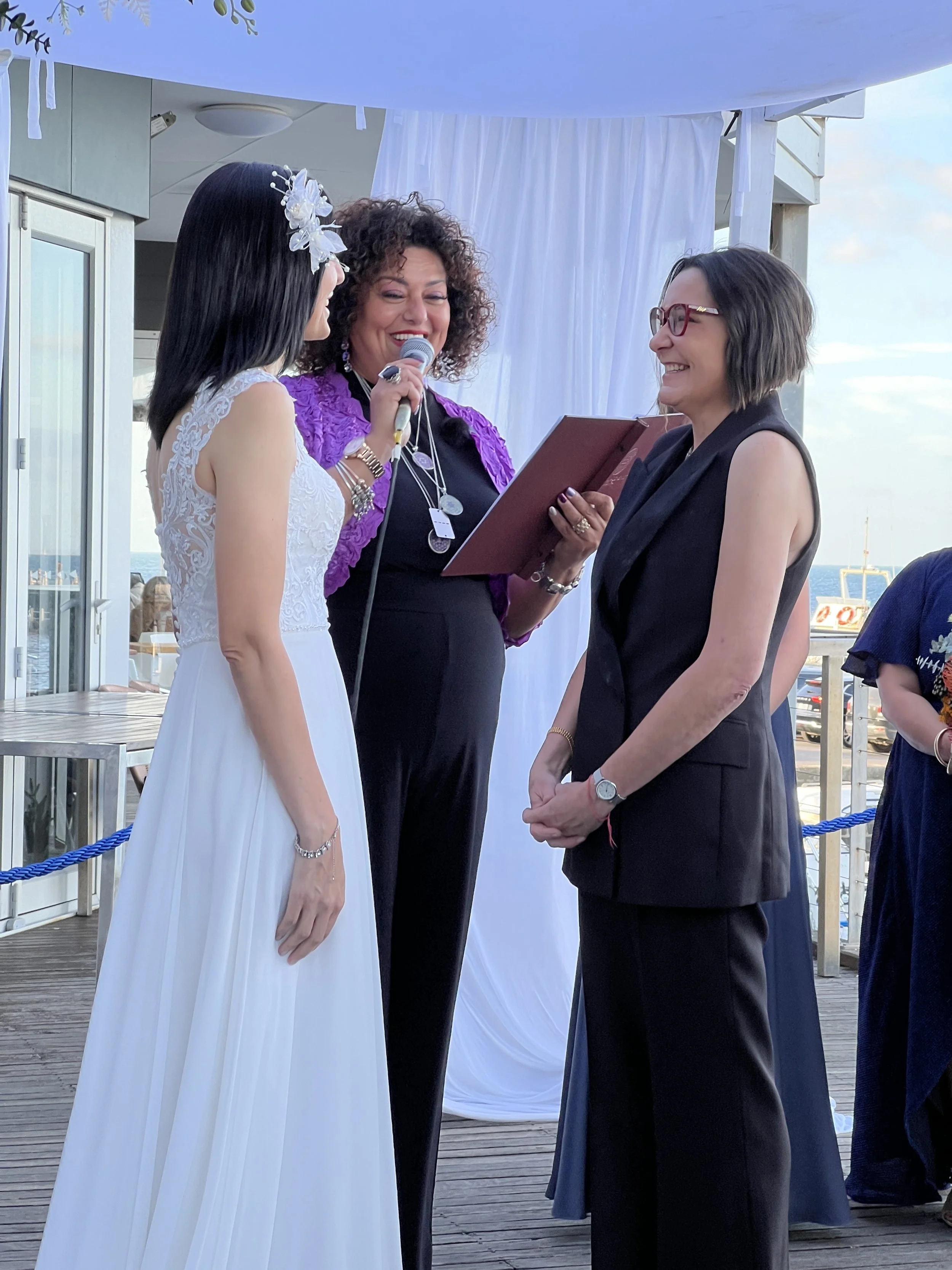A wedding ceremony on a wooden deck with a bride in a white dress, a woman with curly hair reading from a book, and another woman in a black suit with short hair and glasses, outdoors near the ocean.