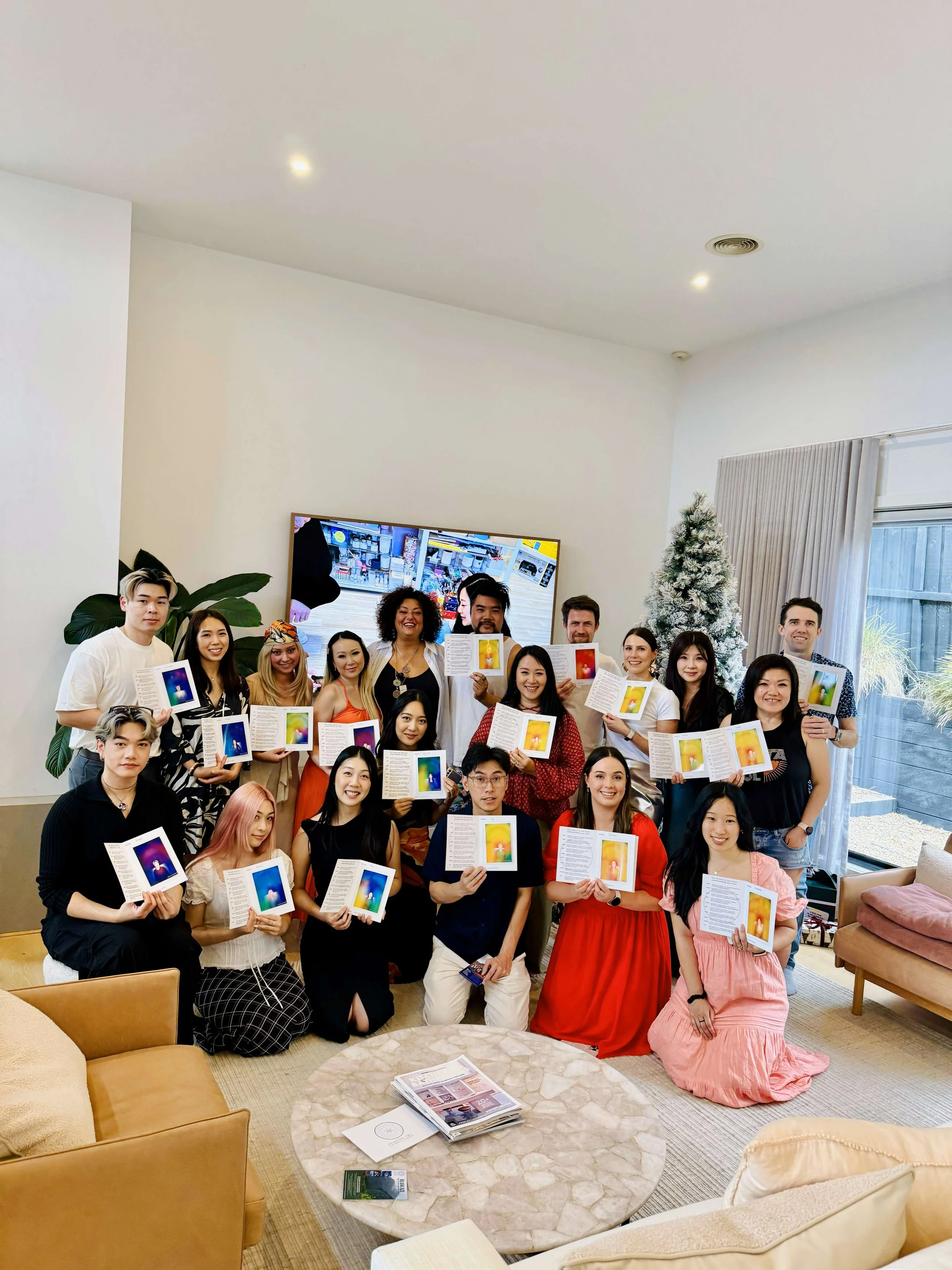 Group of 20 people smiling and holding open books in a living room with a Christmas tree, flat-screen TV, and large window.