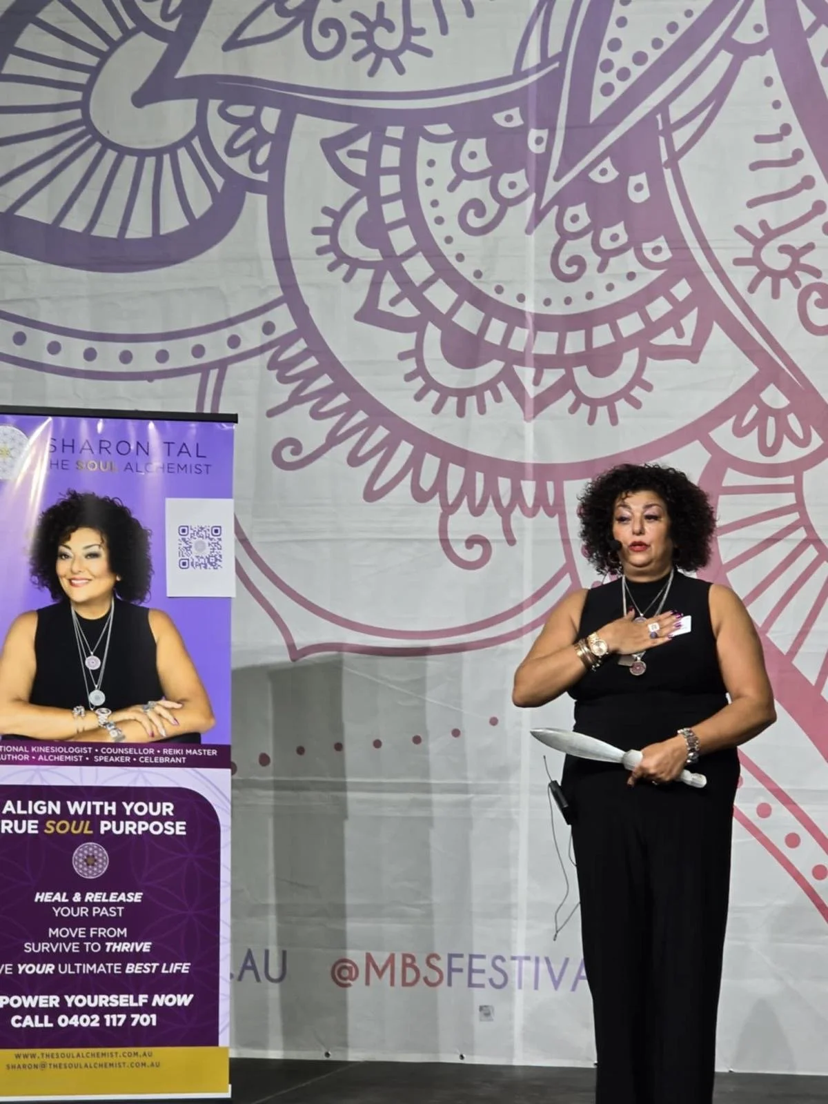 A woman with curly hair in a black sleeveless dress standing on stage, holding a feather and gesturing with her right hand, speaking at an event. Behind her is a large decorative backdrop with purple abstract designs, and to her left, there is a purp