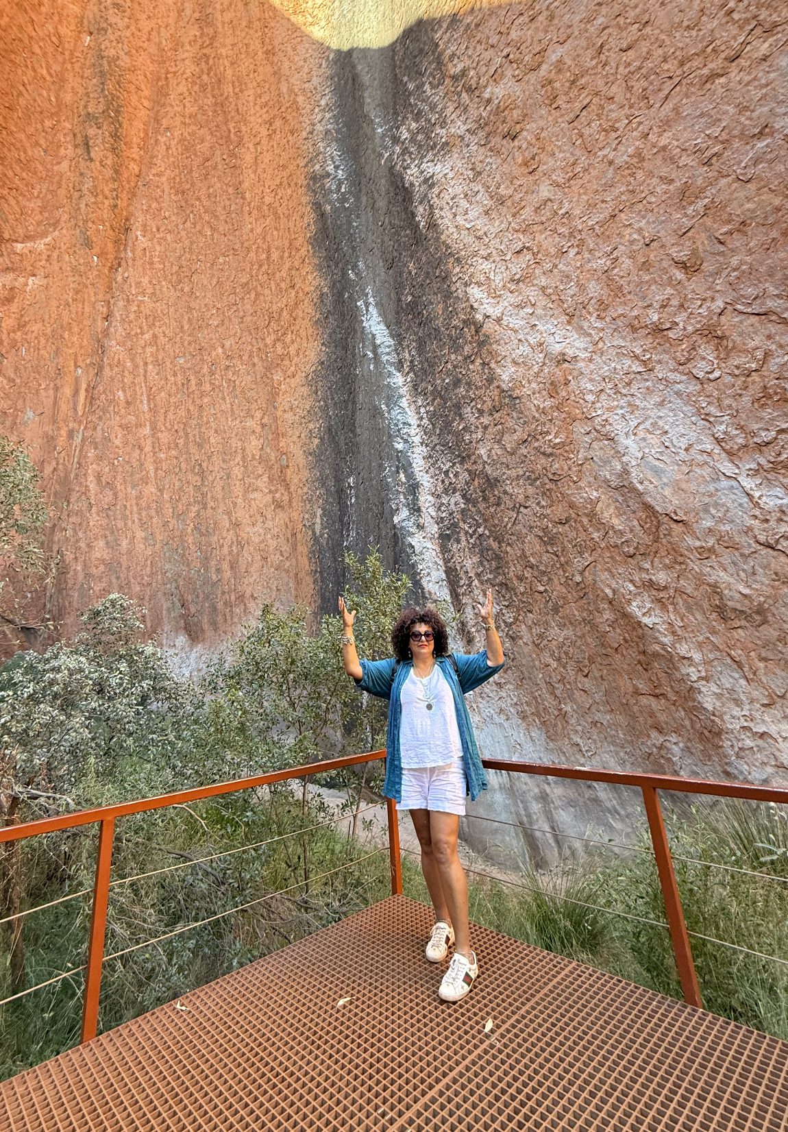 A woman standing on a viewing platform next to a large red rock face with a small waterfall flowing down it.