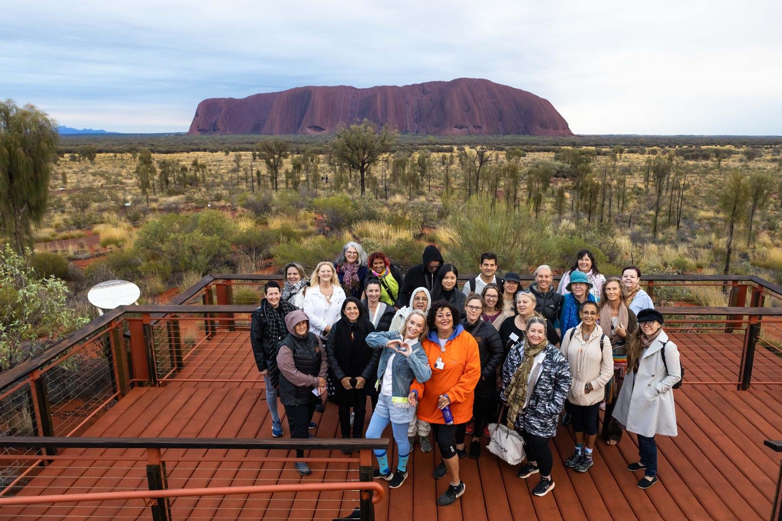 Group of people on a wooden observation deck overlooking a desert landscape with trees and Uluru in the background.