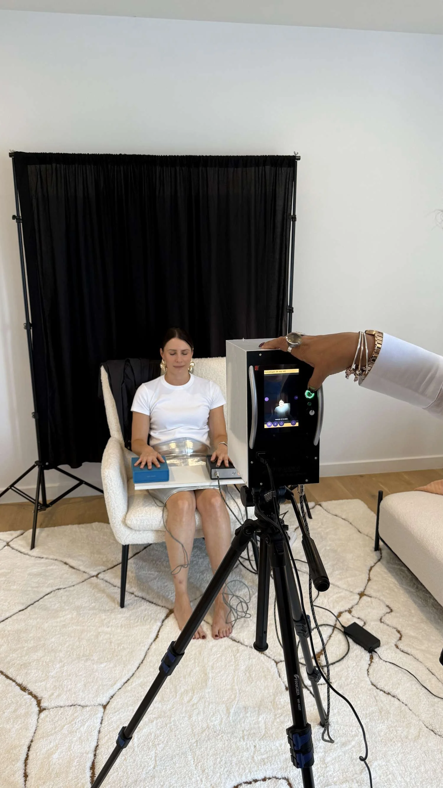 A woman is seated in a chair with a black backdrop behind her. She has her eyes closed and is wearing a white top, a metallic belt, and jewelry. There is camera equipment and a person operating it in front of her, capturing a video or photo.
