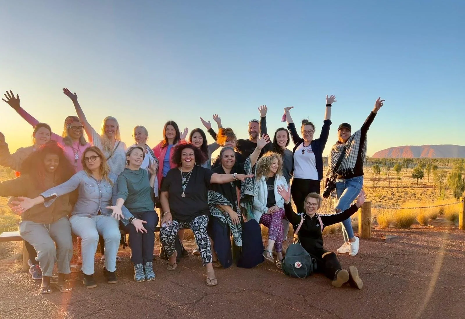 Group of diverse women and men smiling and raising arms in a desert landscape during sunset.